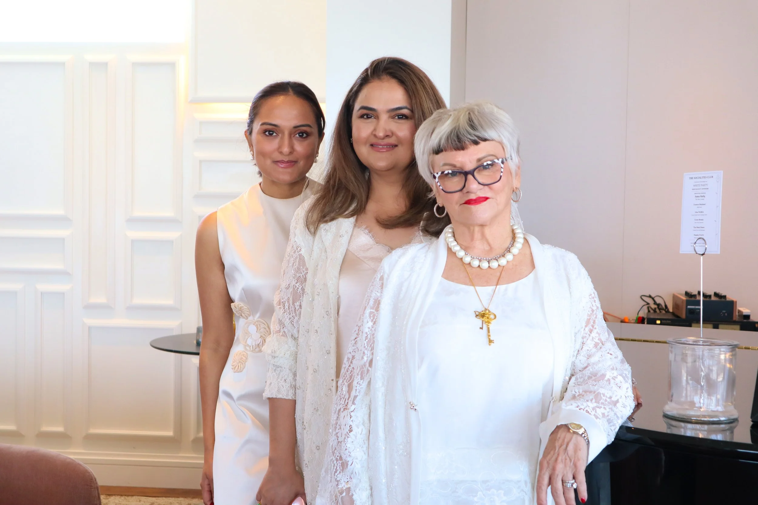 Three women standing together indoors. The woman in front wears glasses, a white lace dress, a pearl necklace, and has short gray hair. The woman in the middle has long brown hair and a light-colored dress. The woman in the back has dark hair, wears 