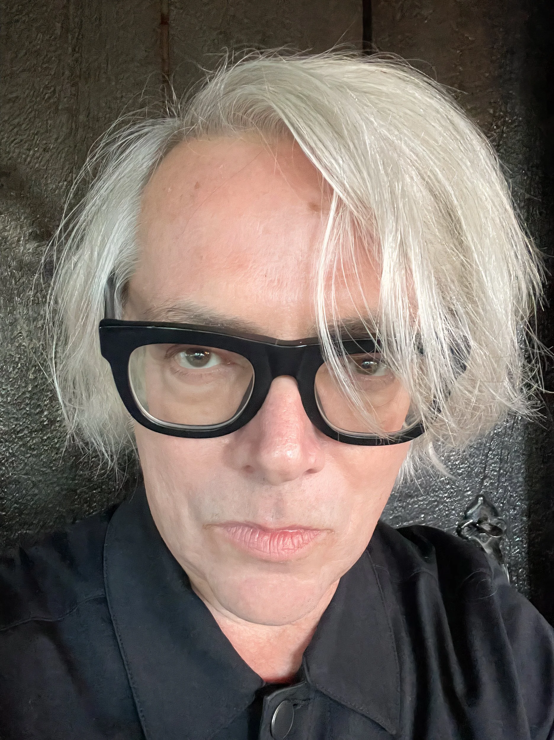 A close-up selfie of a person with light skin, platinum blonde hair, wearing black-rimmed glasses, dressed in black, against a dark background.