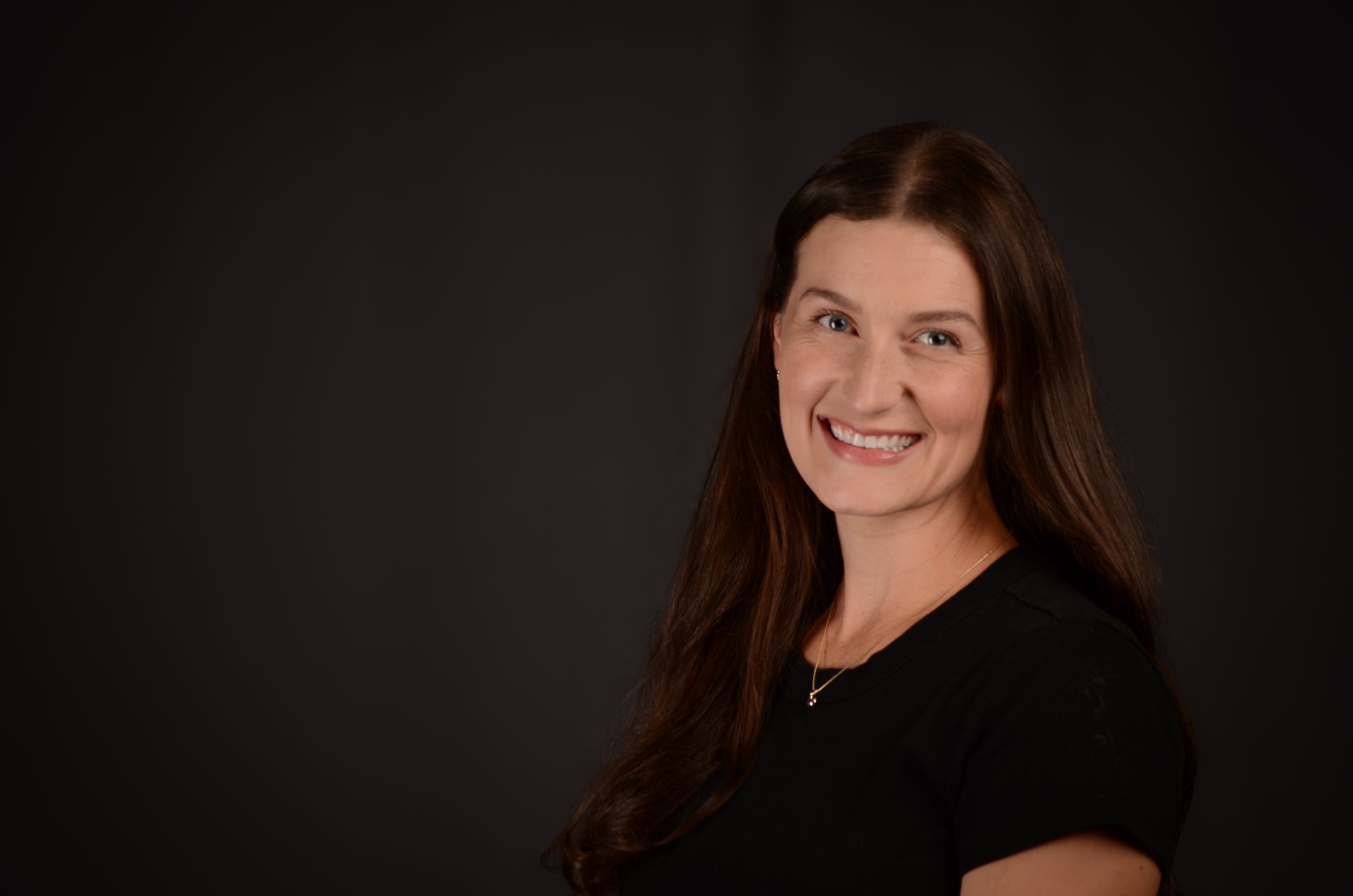 A smiling woman with long brown hair, wearing a black top and a necklace, posed against a dark background.