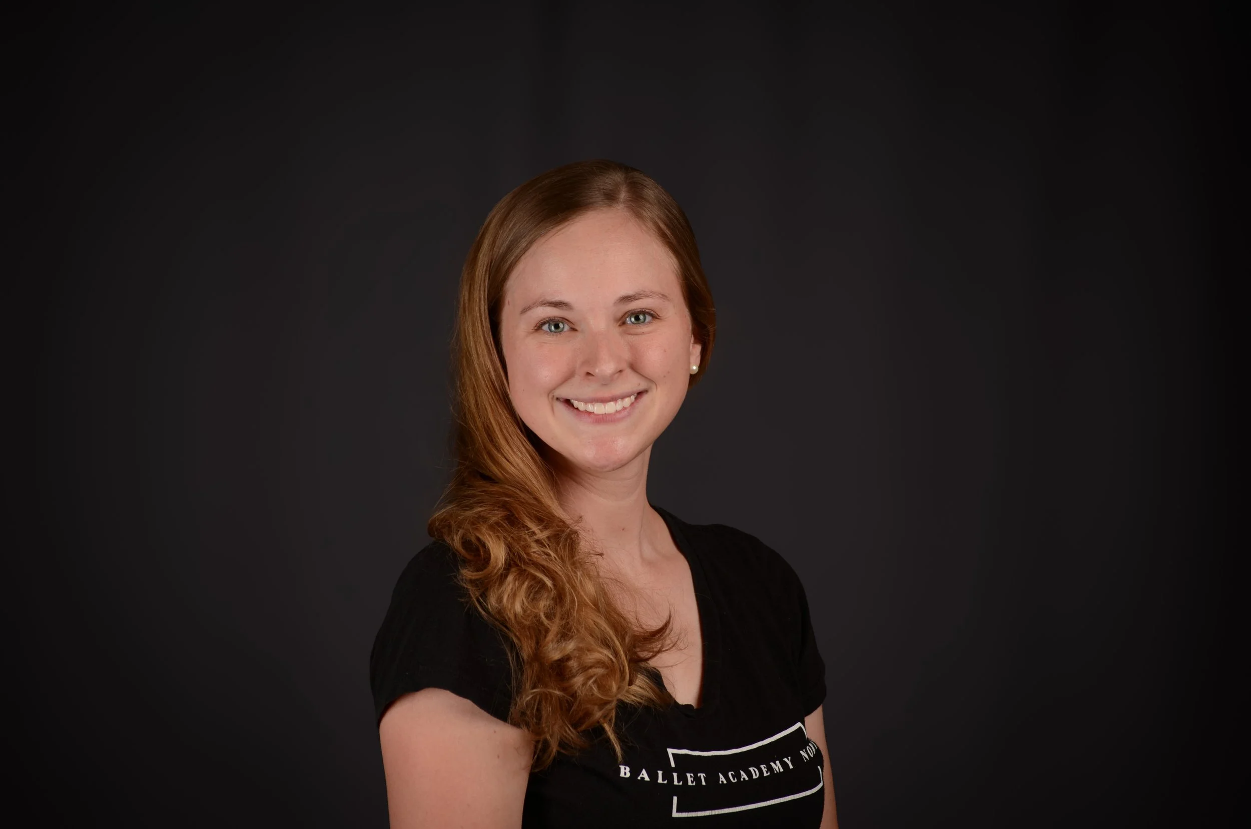 Portrait of a young woman with long red hair, smiling, wearing a black shirt with white text, in front of a dark background.