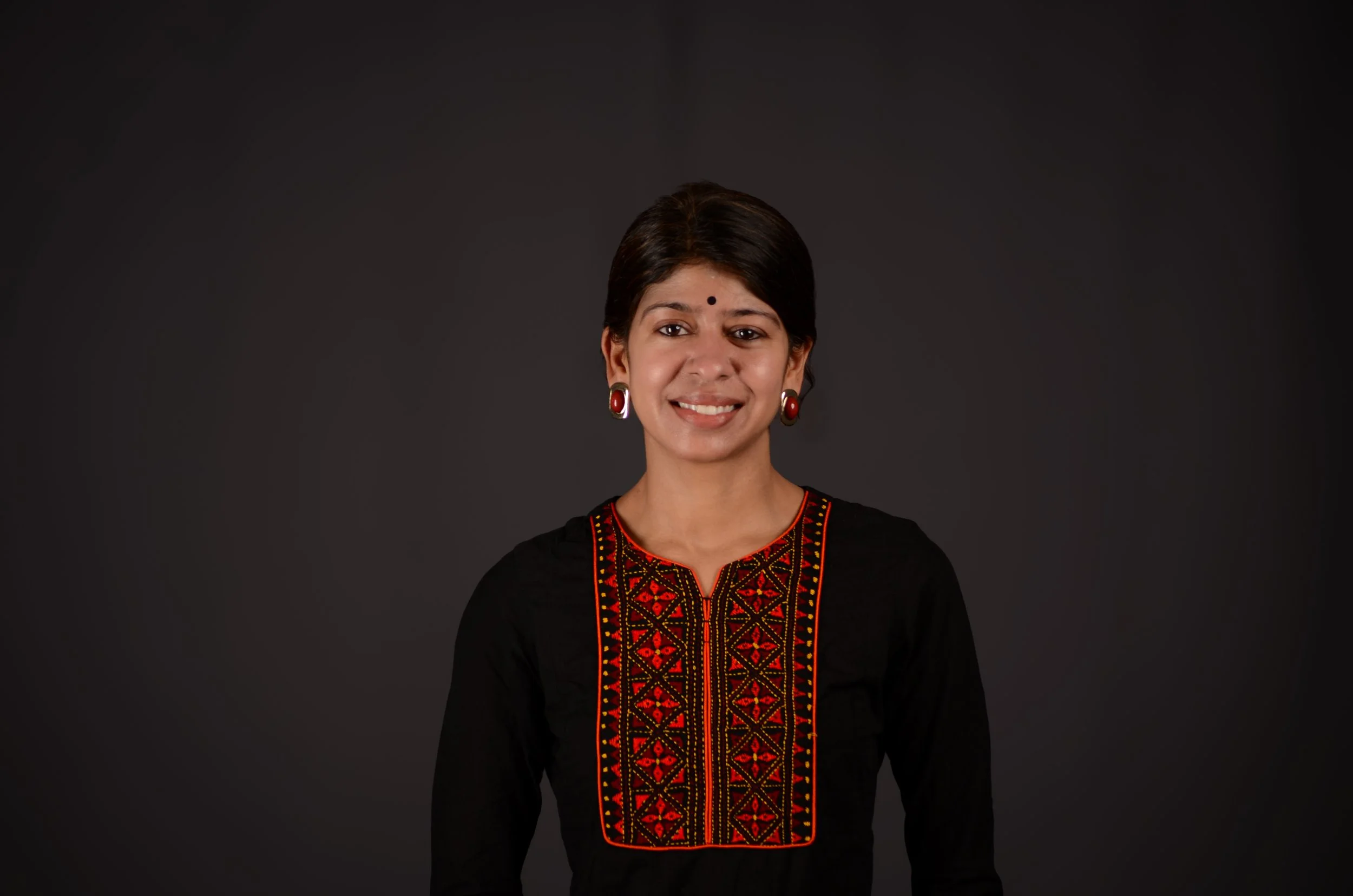 A woman with dark hair wearing traditional Indian attire, including earrings and an embroidered black top, smiling against a plain dark background.