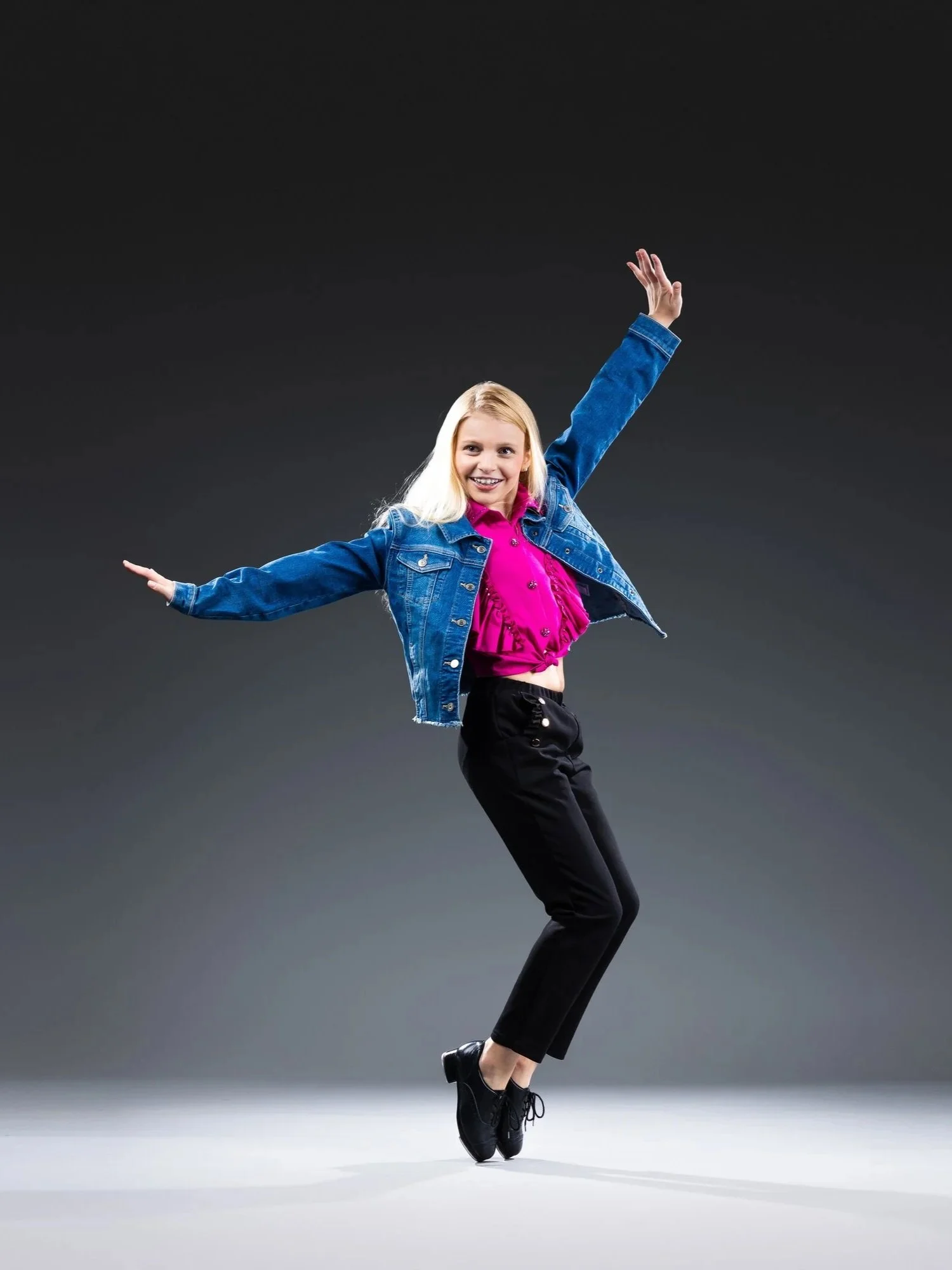 A young girl ice skating in a pose, wearing a denim jacket, pink top, black pants, and black ice skates, against a dark gradient background.