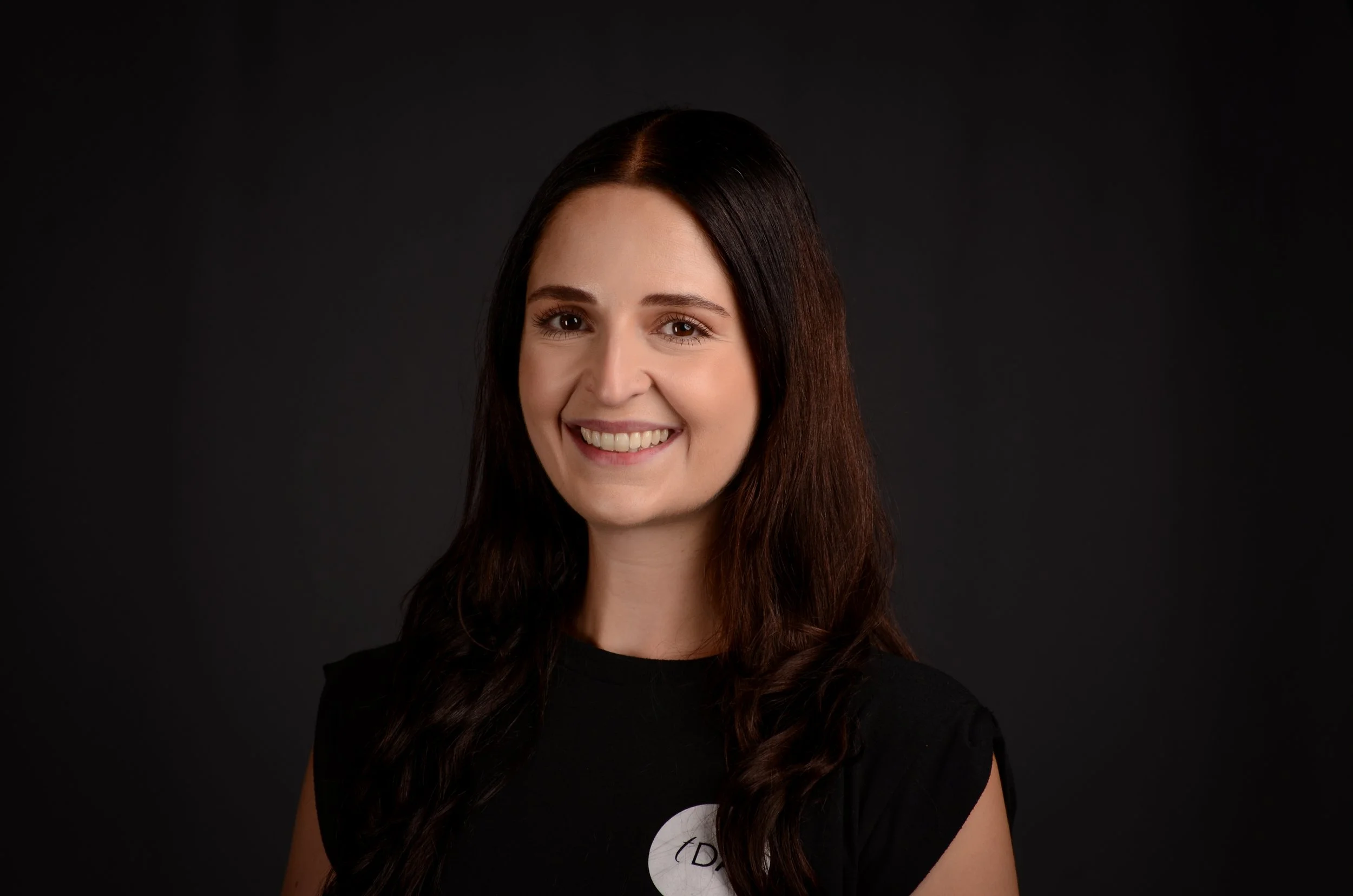 A young woman with long brown hair and brown eyes, smiling, wearing a black top against a dark background.