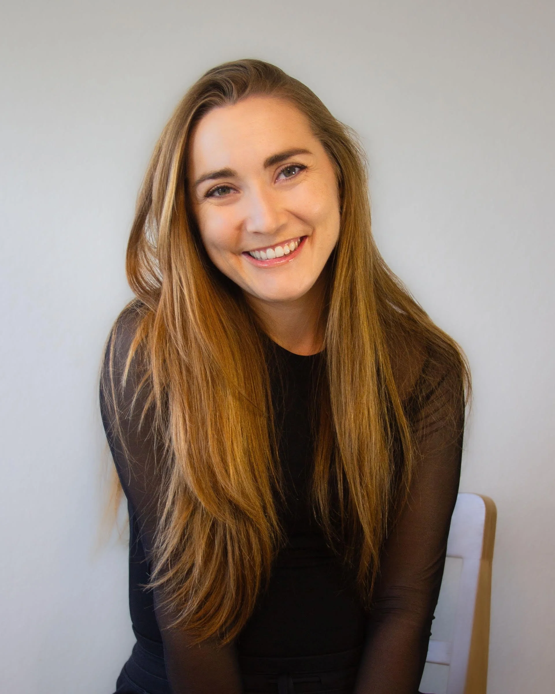 A smiling young woman with long, light brown hair sitting on a chair against a plain white wall.
