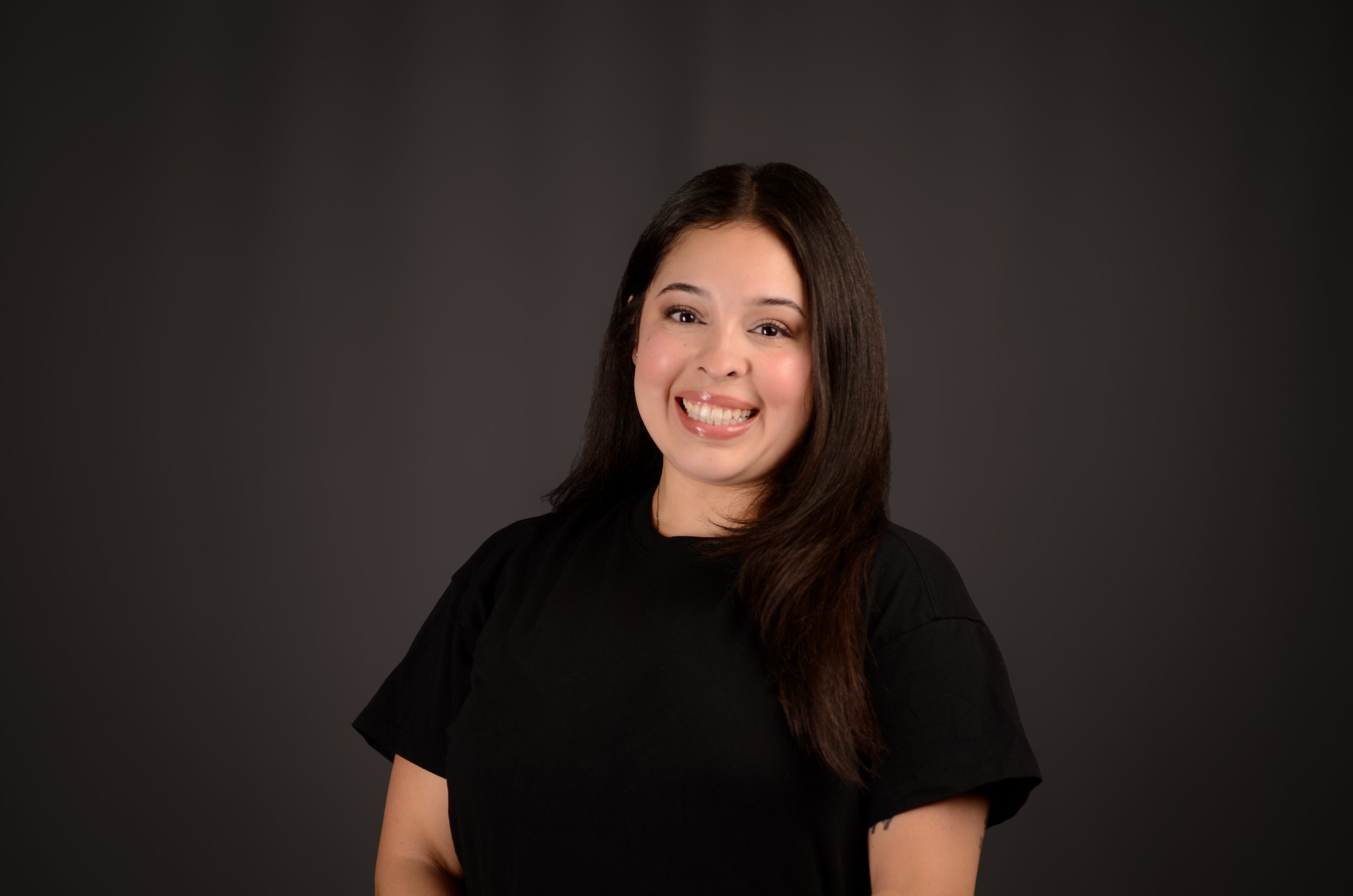 A woman with long dark hair smiling at the camera, wearing a black shirt, against a dark gray background.