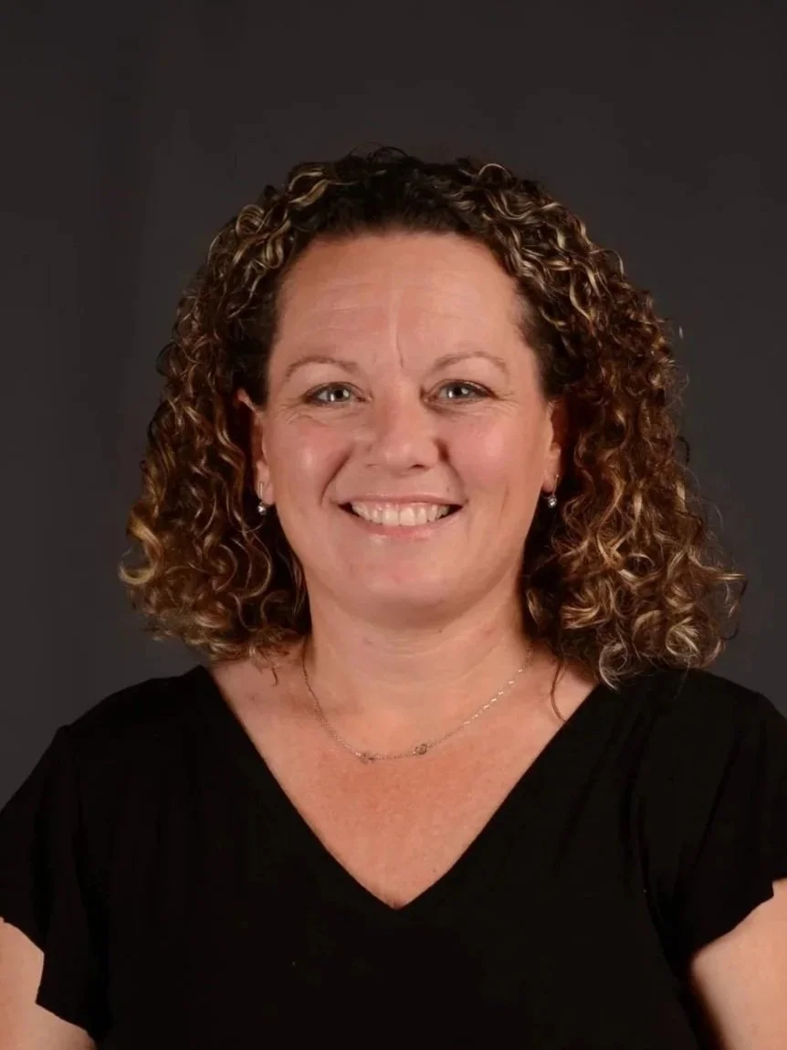 A woman with curly brown hair smiling at the camera, wearing a black top and small earrings against a dark gray background.