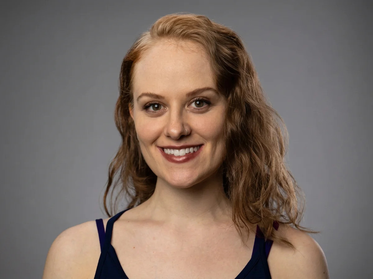 A young woman with red wavy hair smiling at the camera, wearing a dark blue top against a gray background.