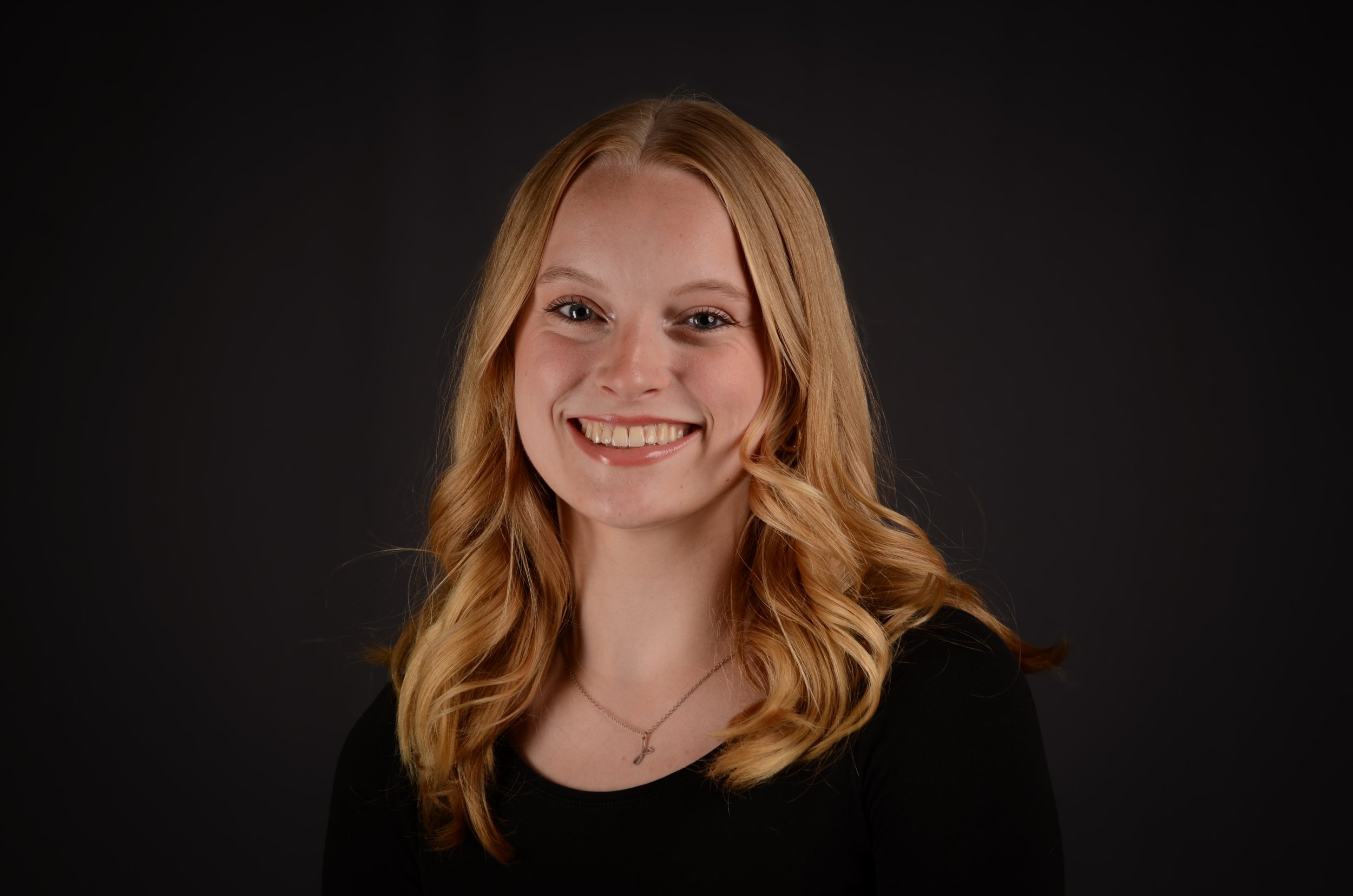 Portrait of a young woman with long, wavy red hair, smiling, wearing a black top, silver necklace, and standing against a dark background.