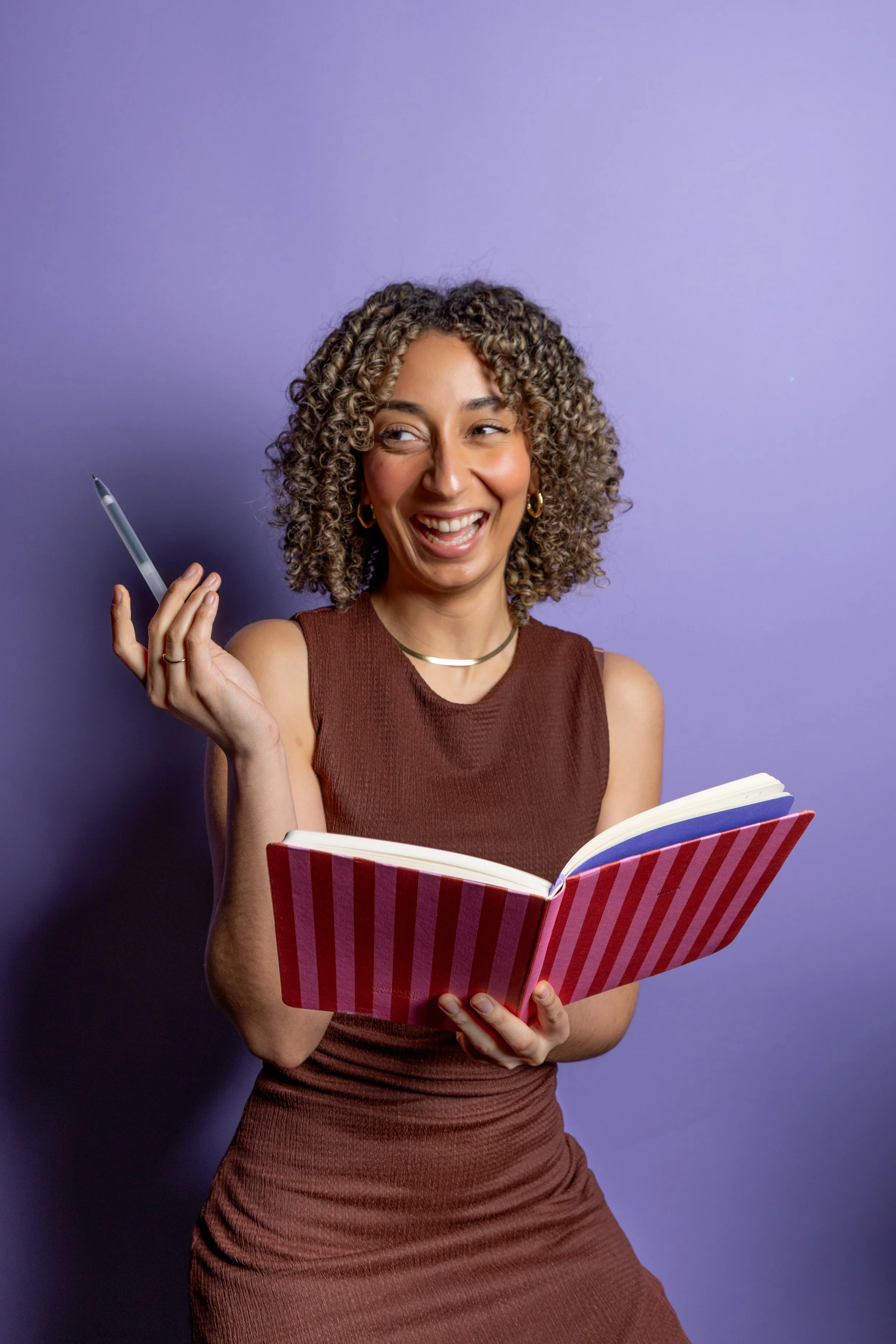 Sierra sits, laughing with her notebook and pen. She is wearing a brown dress before a lavender background