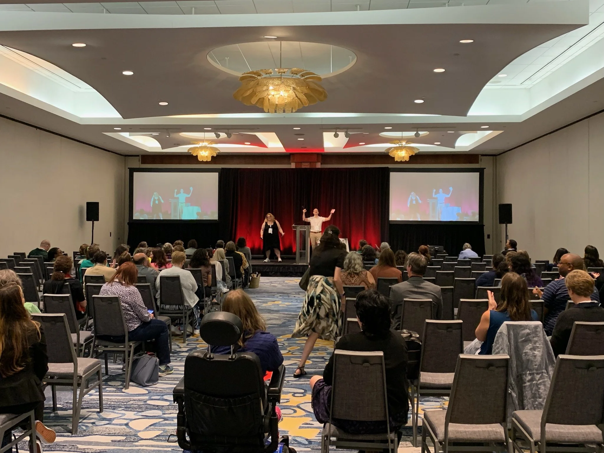 Conference room with a stage, two large screens, and an audience seated in chairs, watching a presentation.