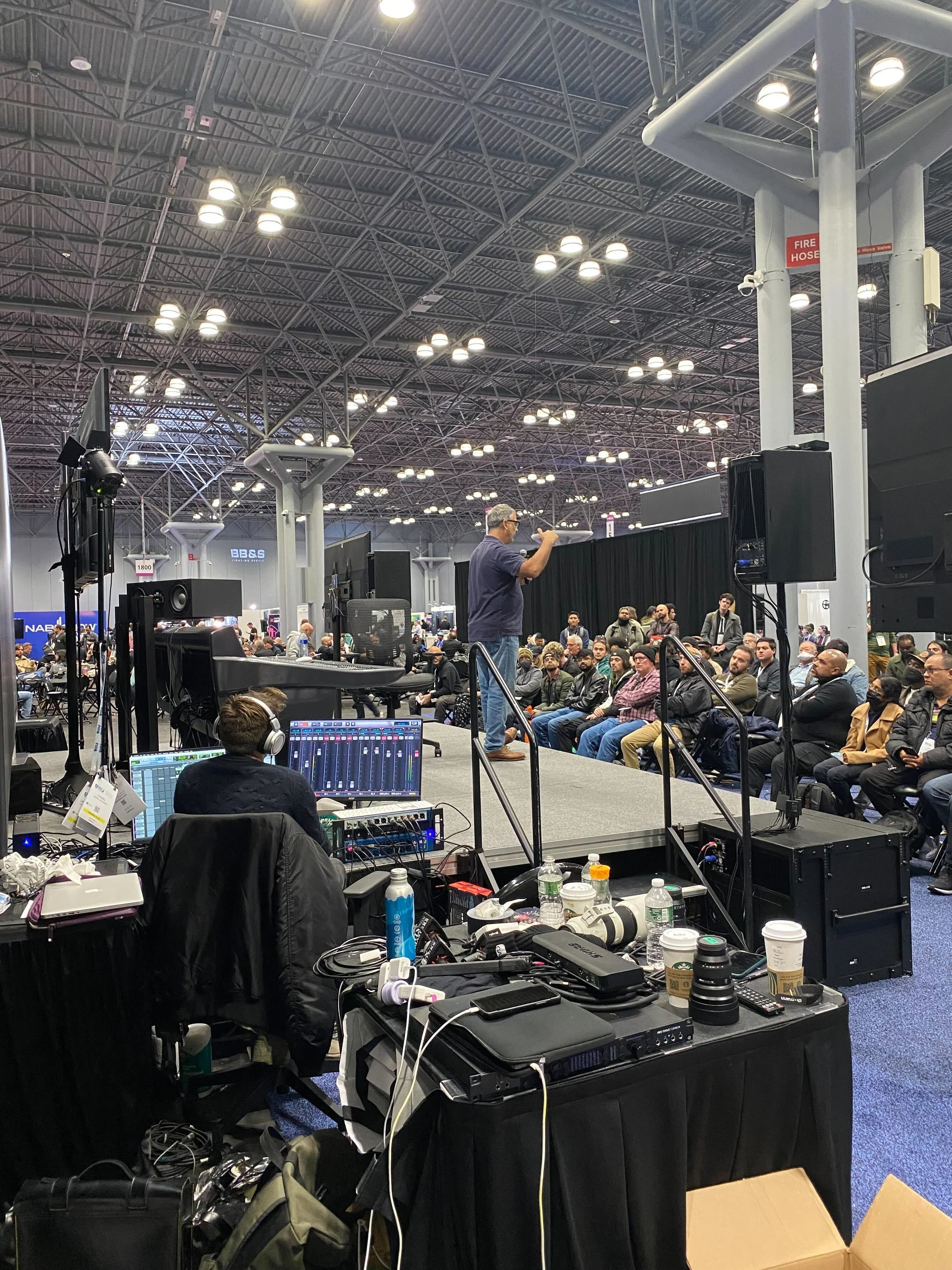 A speaker is giving a presentation on a stage at a conference with an audience of men seated in front. Technical equipment, including monitors, laptops, and audio gear, are visible in the foreground.