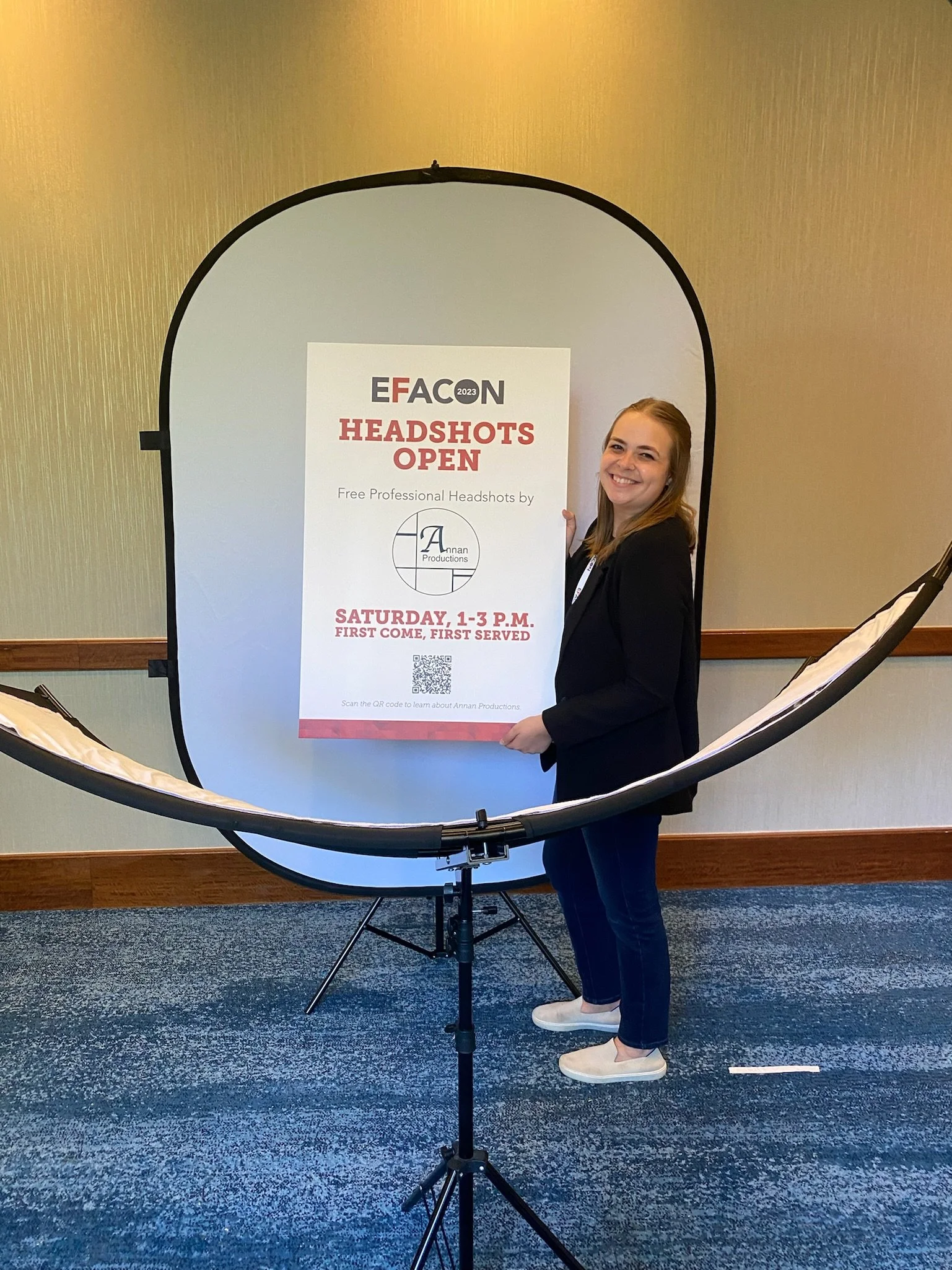 A woman standing next to a sign advertising free professional headshots during an event called EFACON 2023, held Saturday from 1 to 3 p.m. in a room with beige walls and blue carpet.