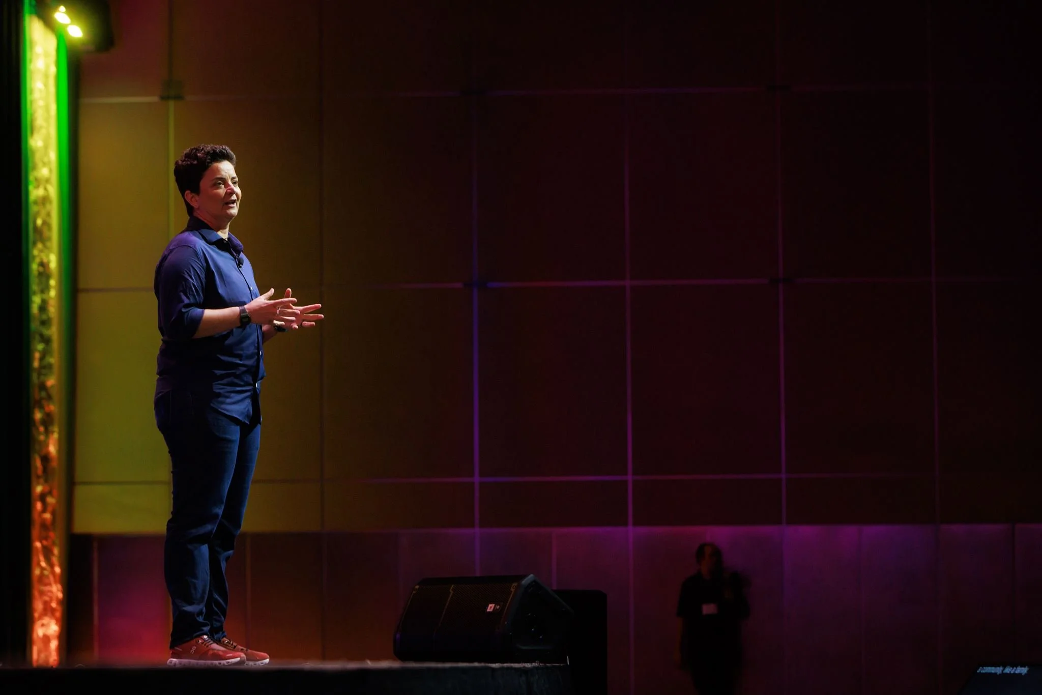 Woman speaking on a stage in a darkened auditorium, illuminated by purple lighting, with a woman in the background.