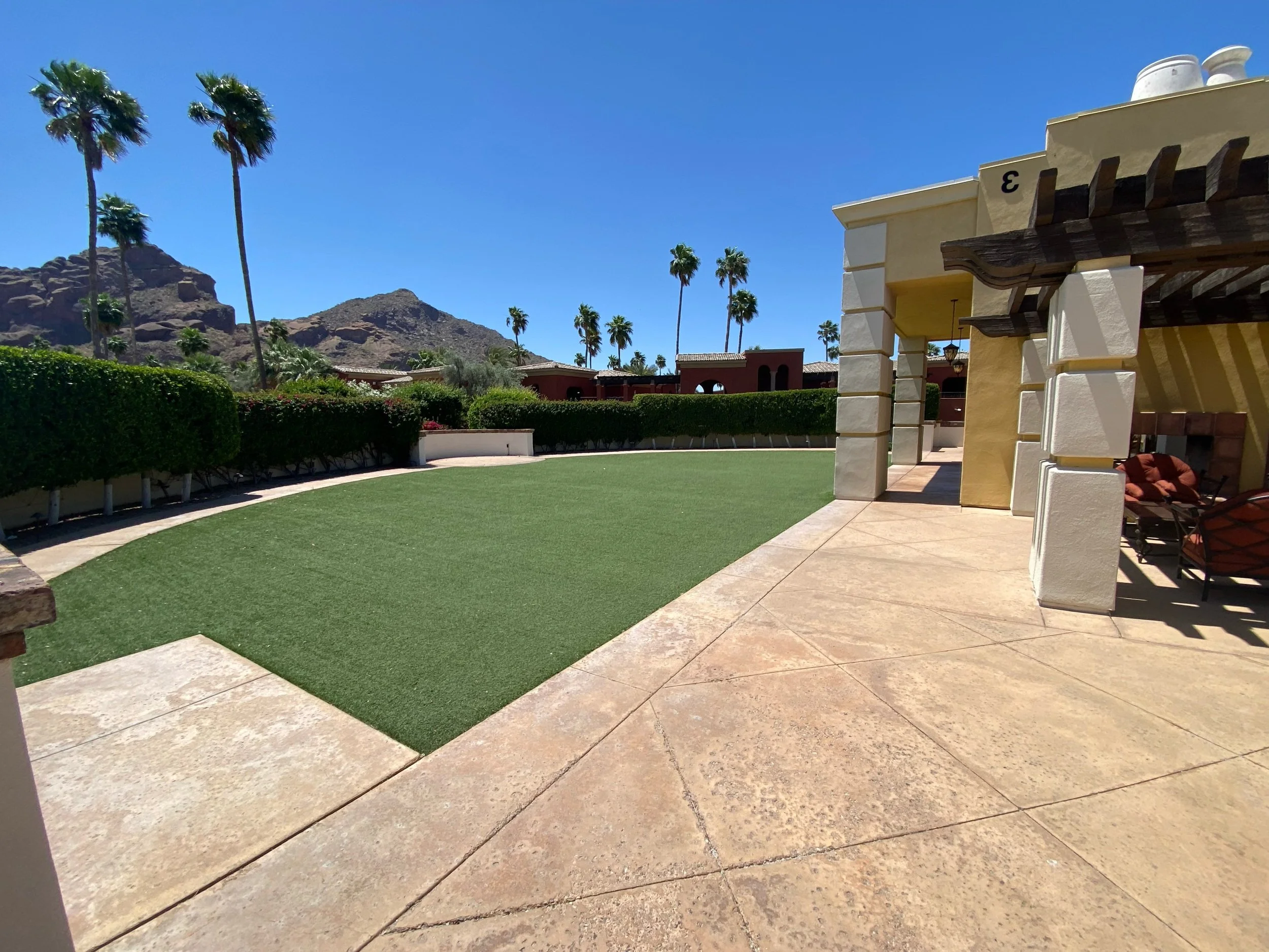 View of a backyard with artificial grass, stone patio, and yellow house with covered patio and outdoor seating. Palm trees and rocky mountains in the background under a clear blue sky.