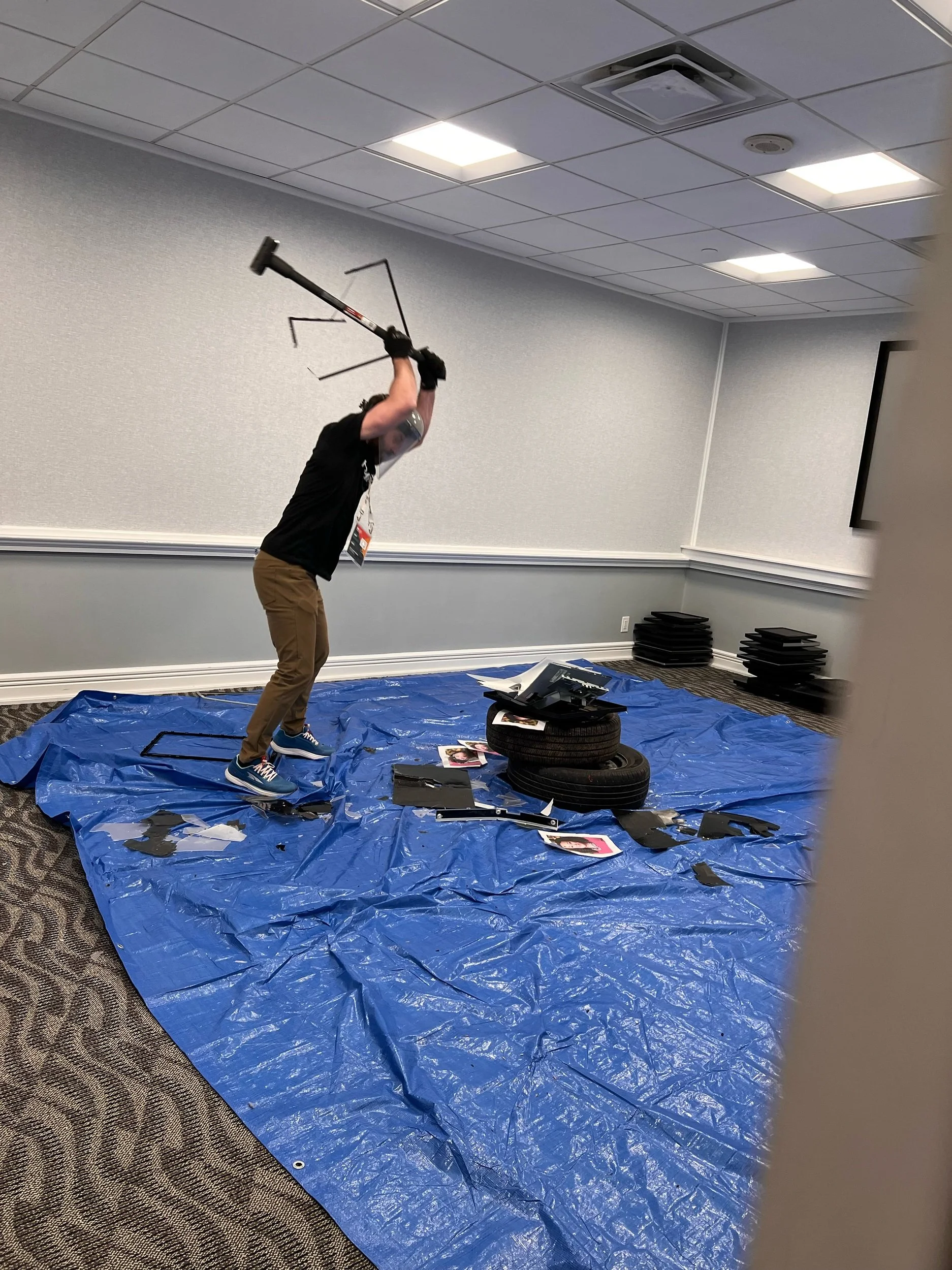 A person in a black shirt and tan pants smashing a television with a crowbar in a conference room, with debris and tires on the floor, covered by a blue tarp.