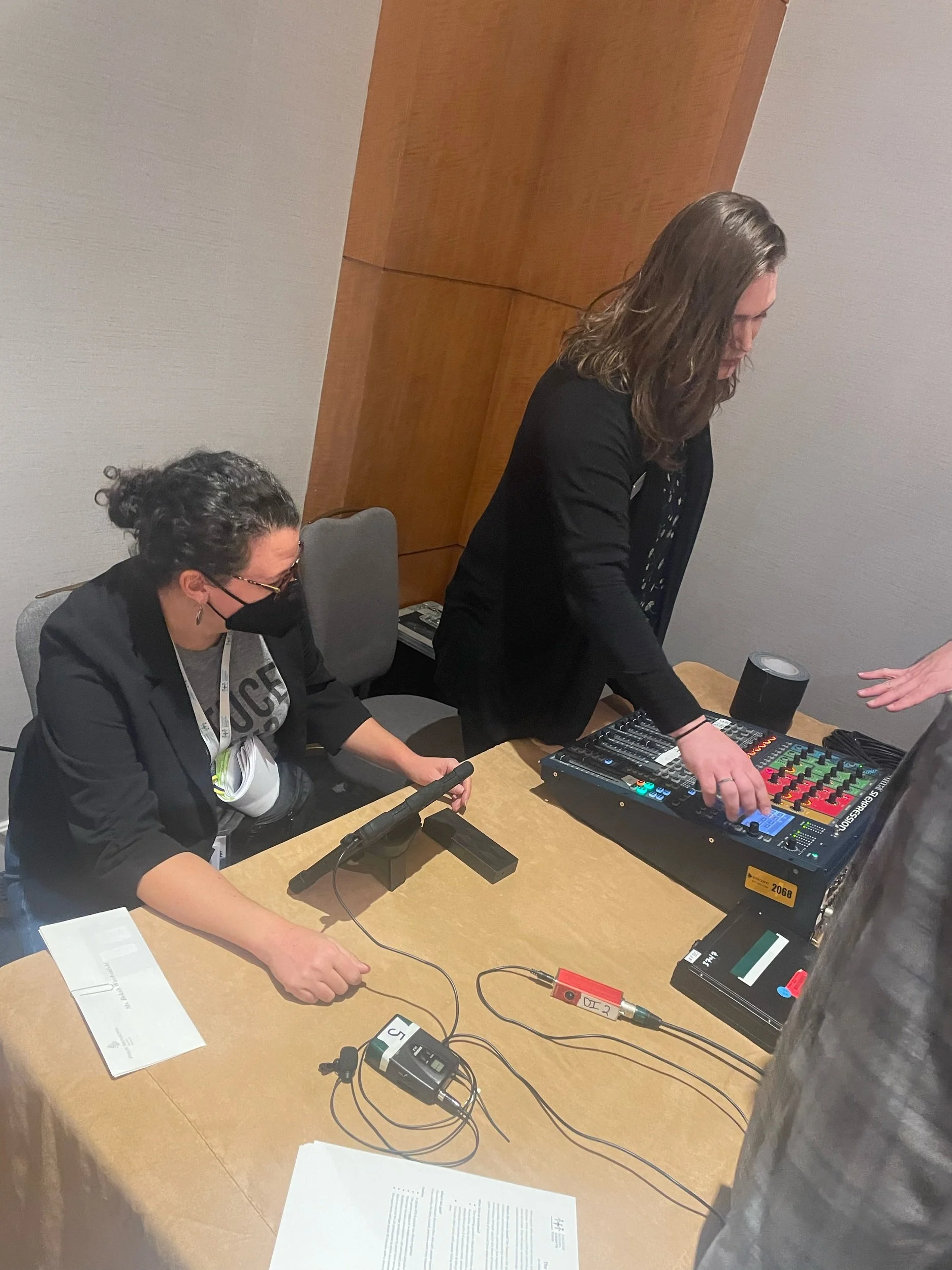 Two women working with audio equipment at a table, one is standing adjusting controls on a sound mixer, and the other is sitting, operating a microphone and connecting wires, both wearing black.
