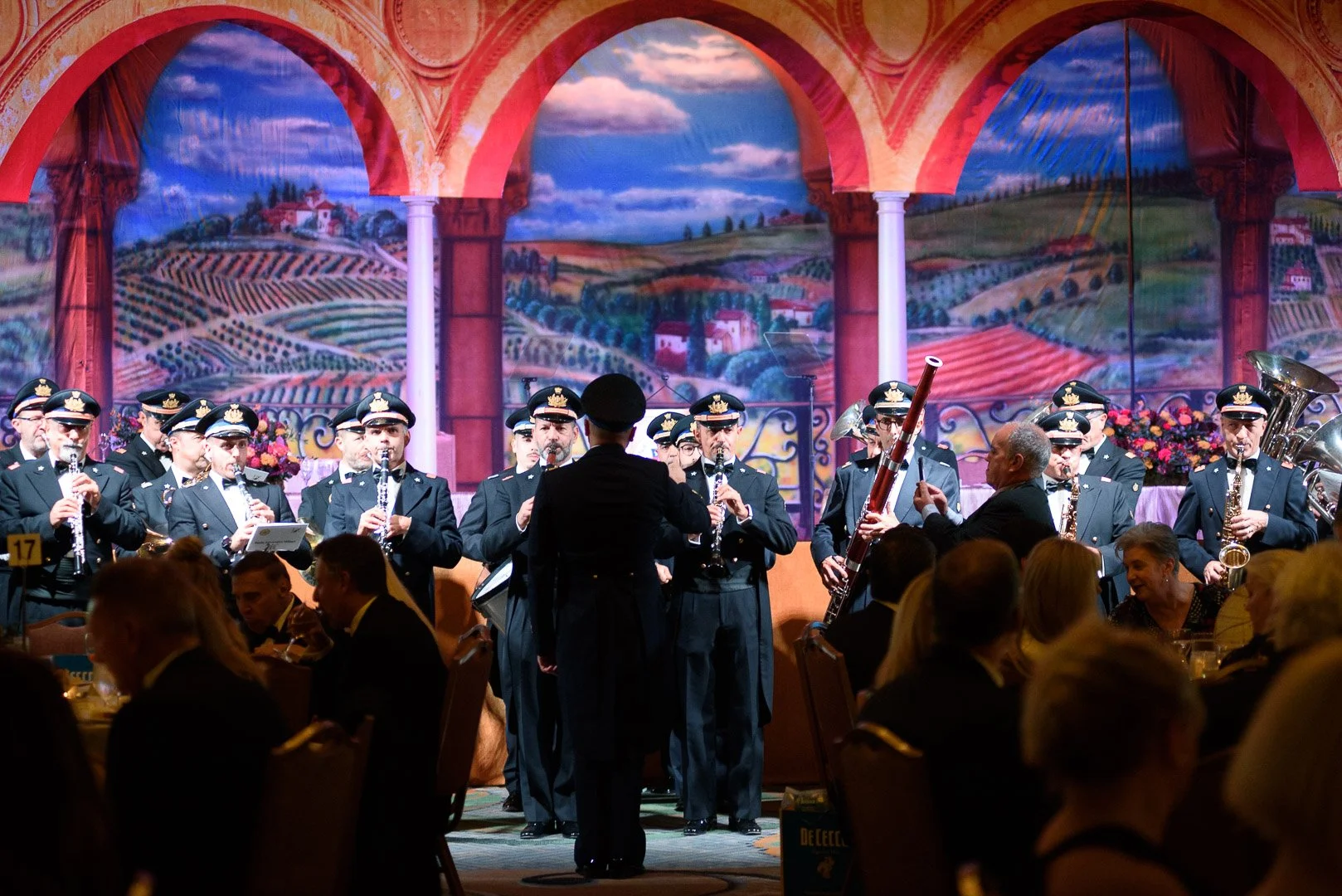 A large group of musicians in police uniforms performing on stage with a painted scenic backdrop of countryside landscapes, while seated guests watch the performance in a banquet setting.