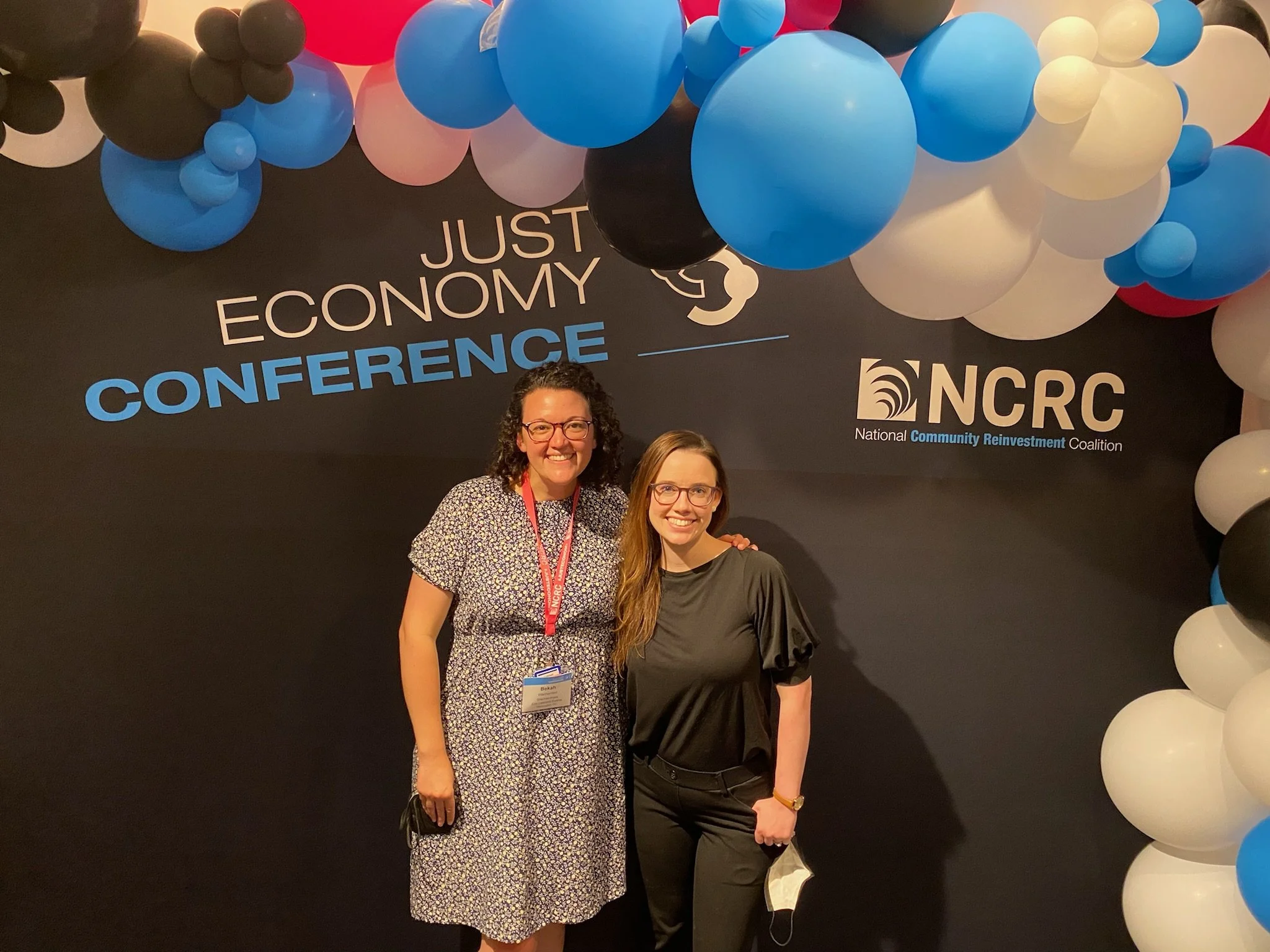 Two women smiling and standing in front of a black backdrop with blue, white, black, and pink balloons arranged in an arch. The backdrop displays the words 'Just Economy Conference' and the logo for the National Community Reinvestment Coalition (NCRC
