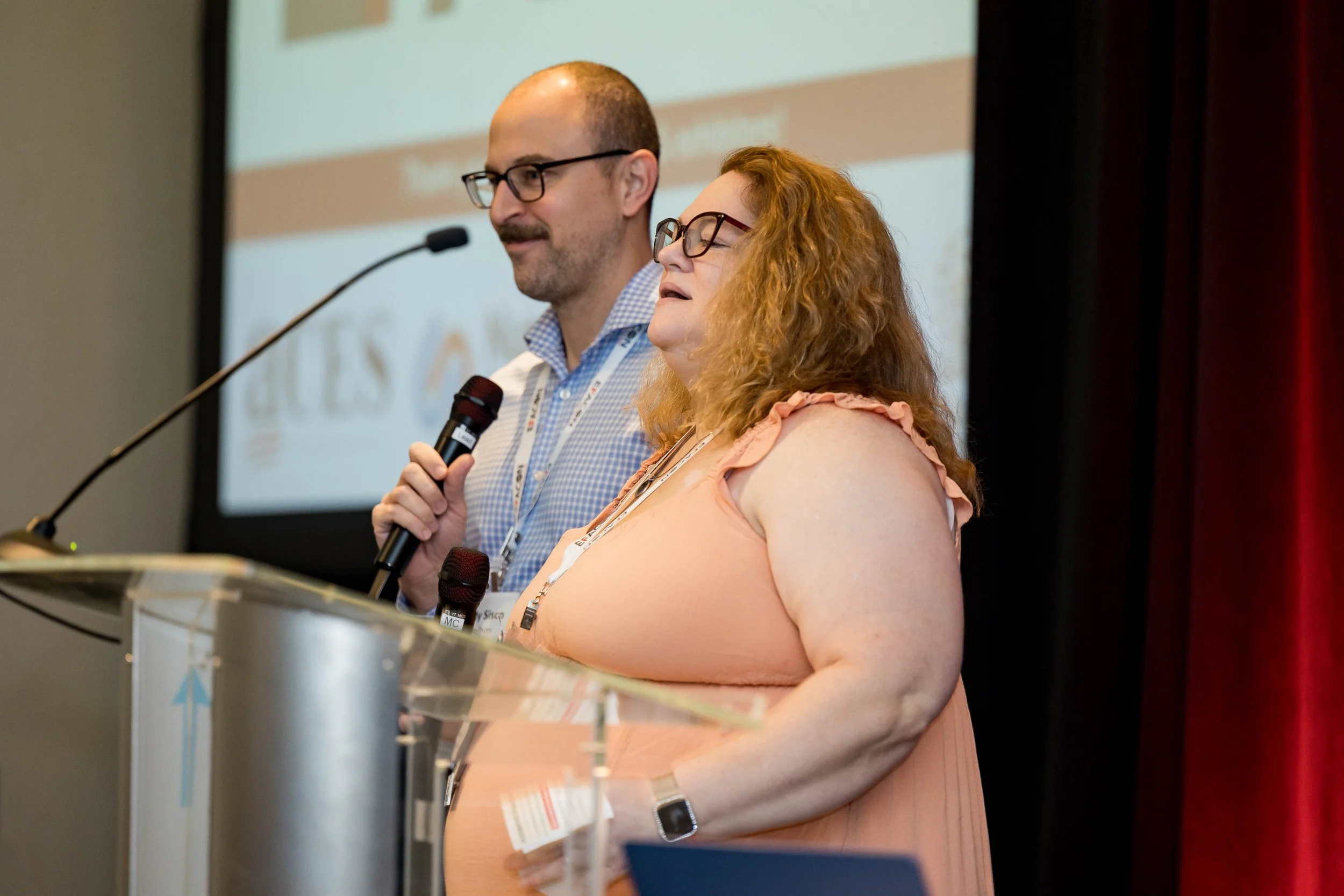 A man and a woman speaking at a conference, standing behind a podium with microphones. The man is wearing glasses and holding a microphone, the woman has red hair and is wearing glasses and a peach dress. They are on a stage with a presentation scree