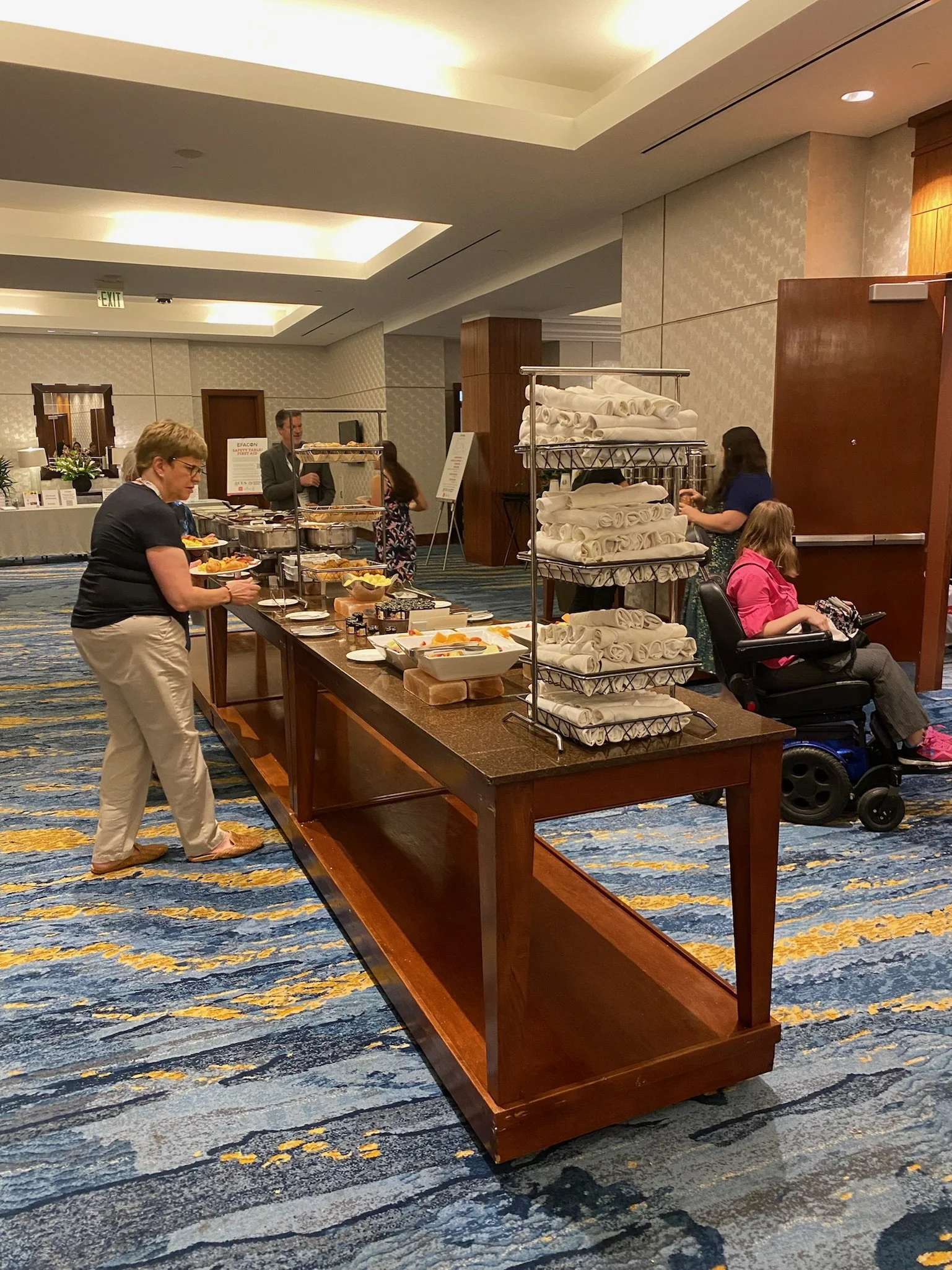 Buffet table with various food items in a conference room setting, with people serving themselves and a woman in a wheelchair nearby.