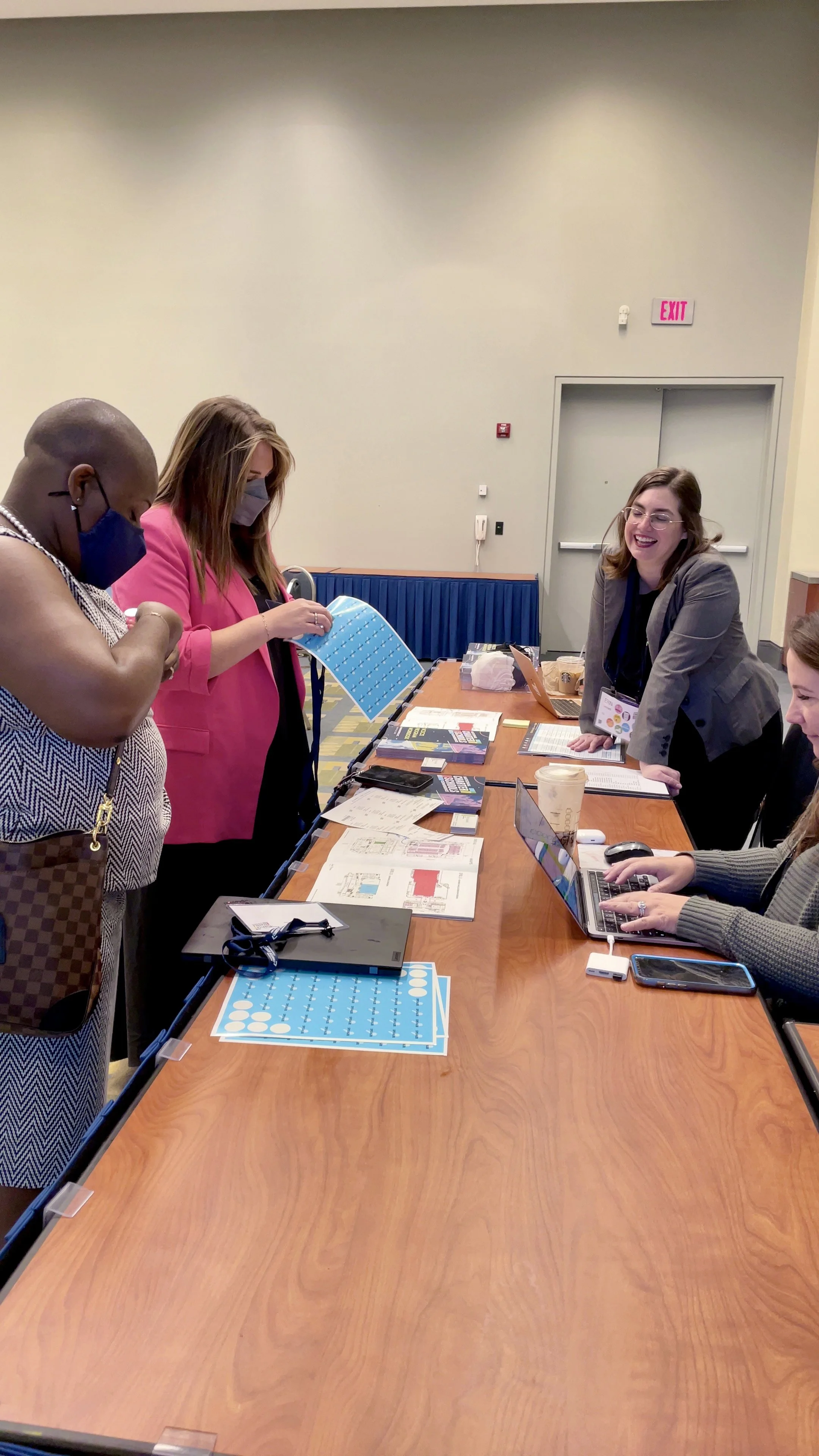 Four women gathered around a conference table engaged in discussion, with laptops, documents, and office supplies on the table, in a conference room.