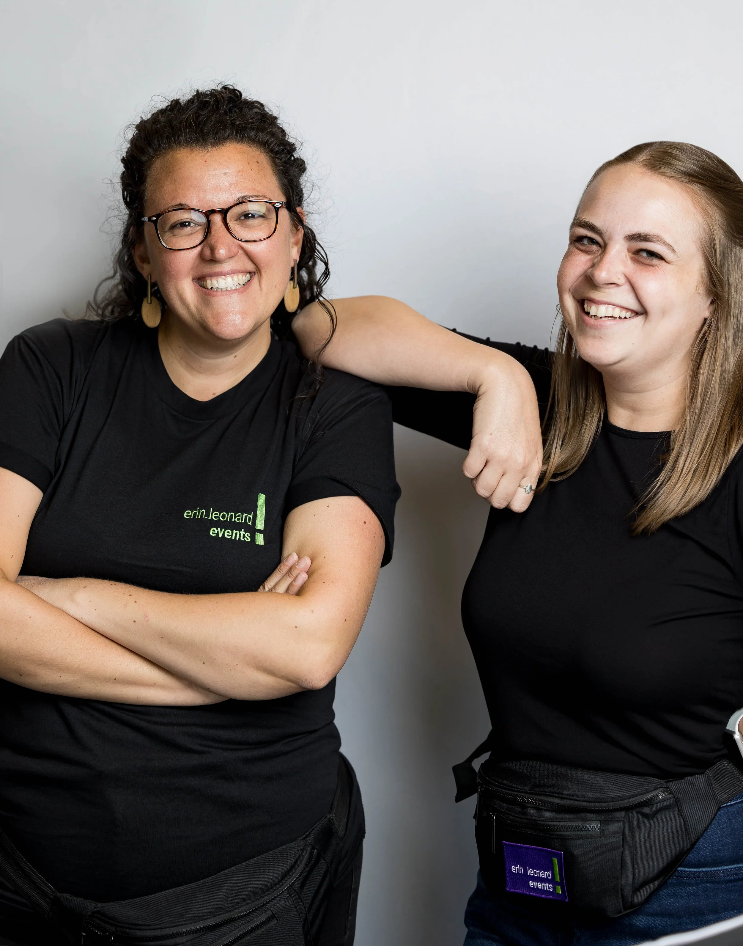 Two women smiling, one with curly hair and glasses, and the other with straight blonde hair, standing against a plain background, wearing black shirts and waist bags, with one woman resting her arm on the other's shoulder.