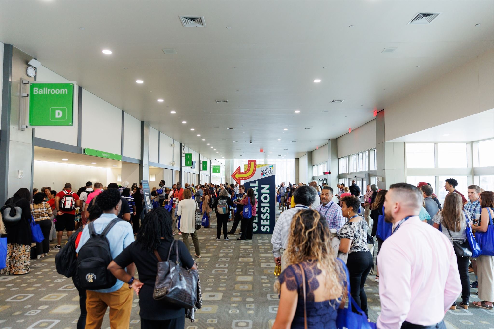 Crowd of people gathering in a large indoor convention center with signs indicating various session areas and an arrow pointing left.