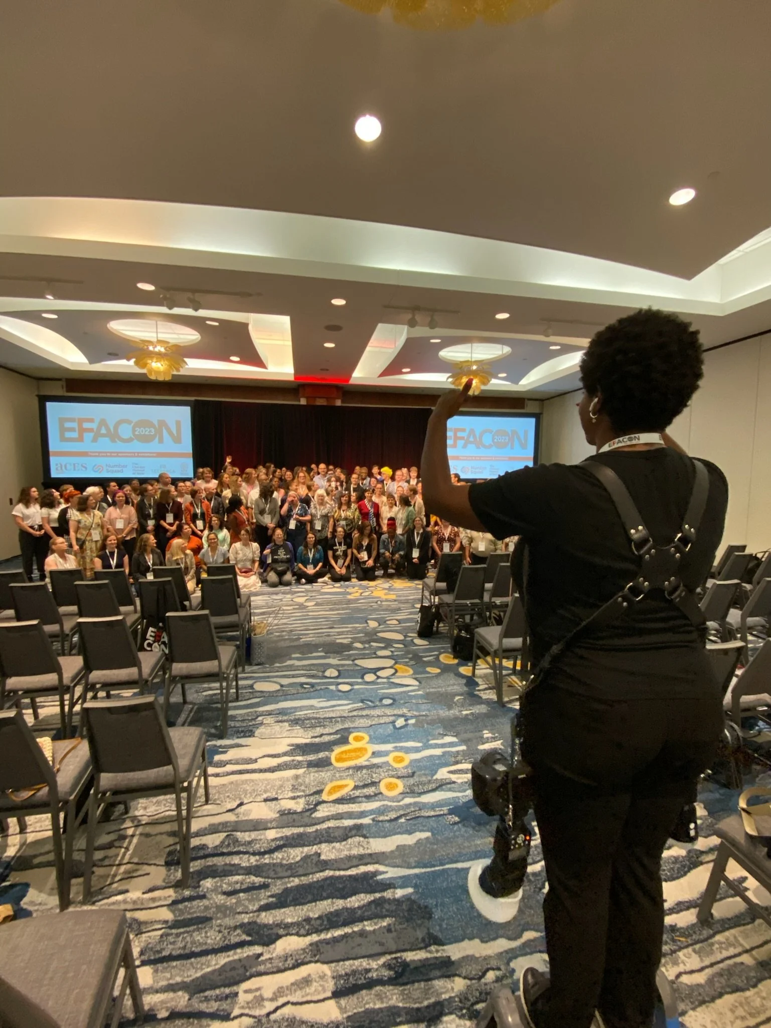 A woman standing in front of a large group of people at a conference, speaking or gesturing towards the audience. The event appears to be the EFACON 2023 conference, as indicated by the screens behind the stage.