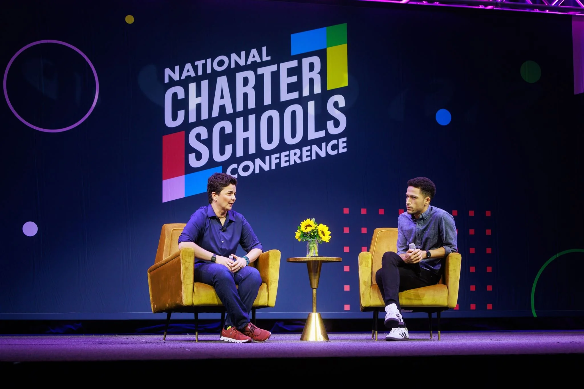 Two people sitting on yellow chairs on a stage at the National Charter Schools Conference, having a conversation. There is a table with a bouquet of sunflowers between them, and a large screen behind displaying the conference logo.