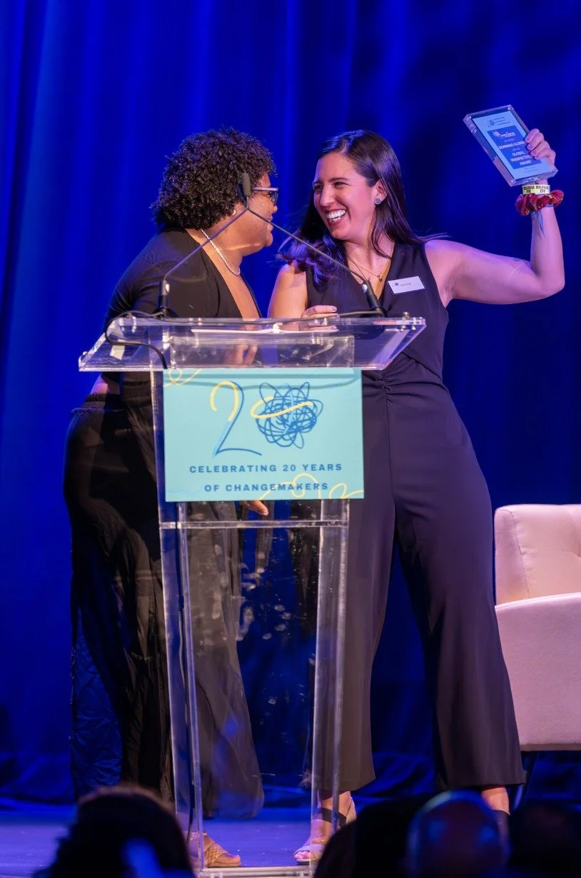 Two women smiling and facing each other on stage, one holding an award, with a blue curtain background and a clear podium with a sign celebrating 20 years of changemakers.