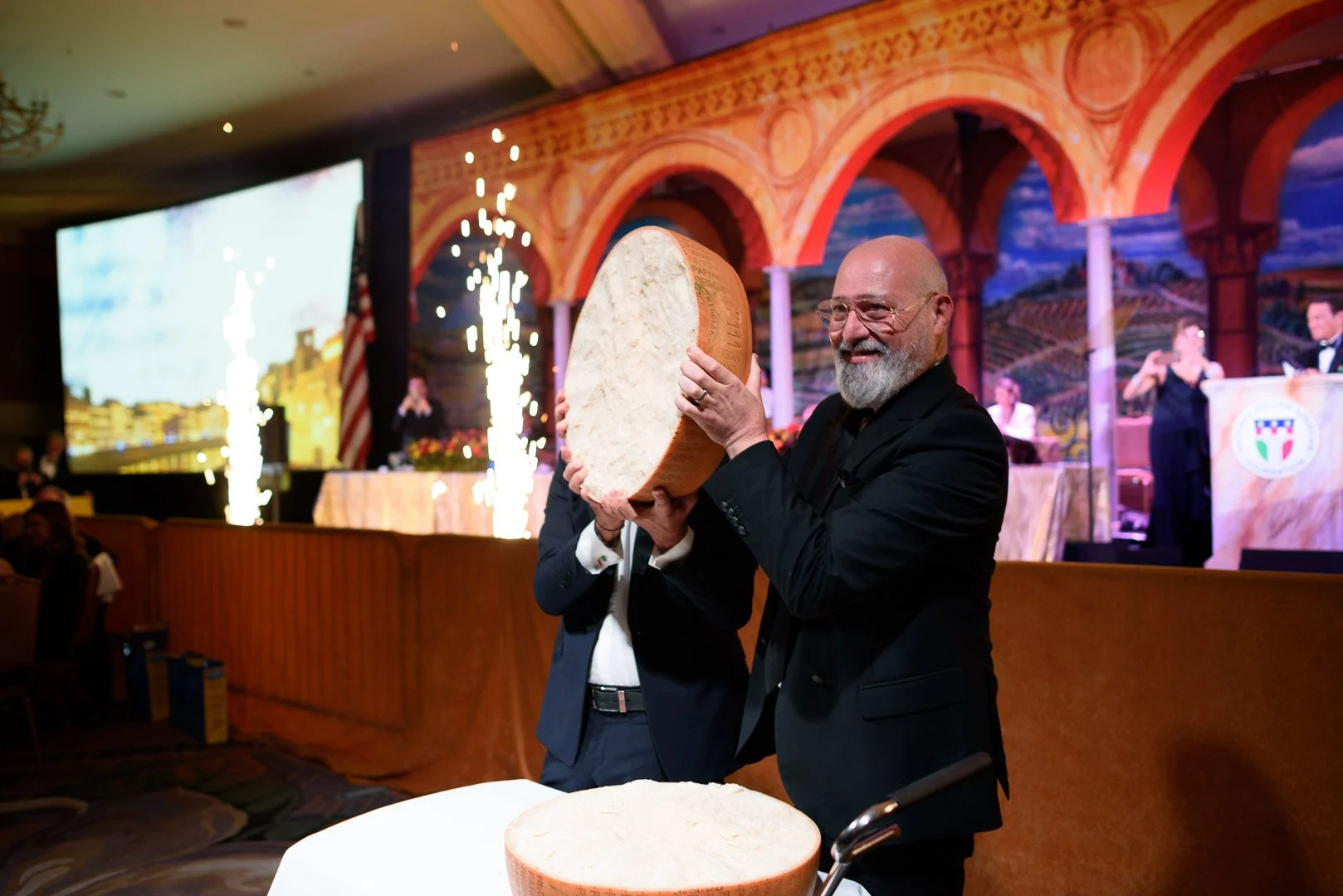 A man holding a large wheel of Parmigiano-Reggiano cheese during a celebration or event, with a stage and band in the background.