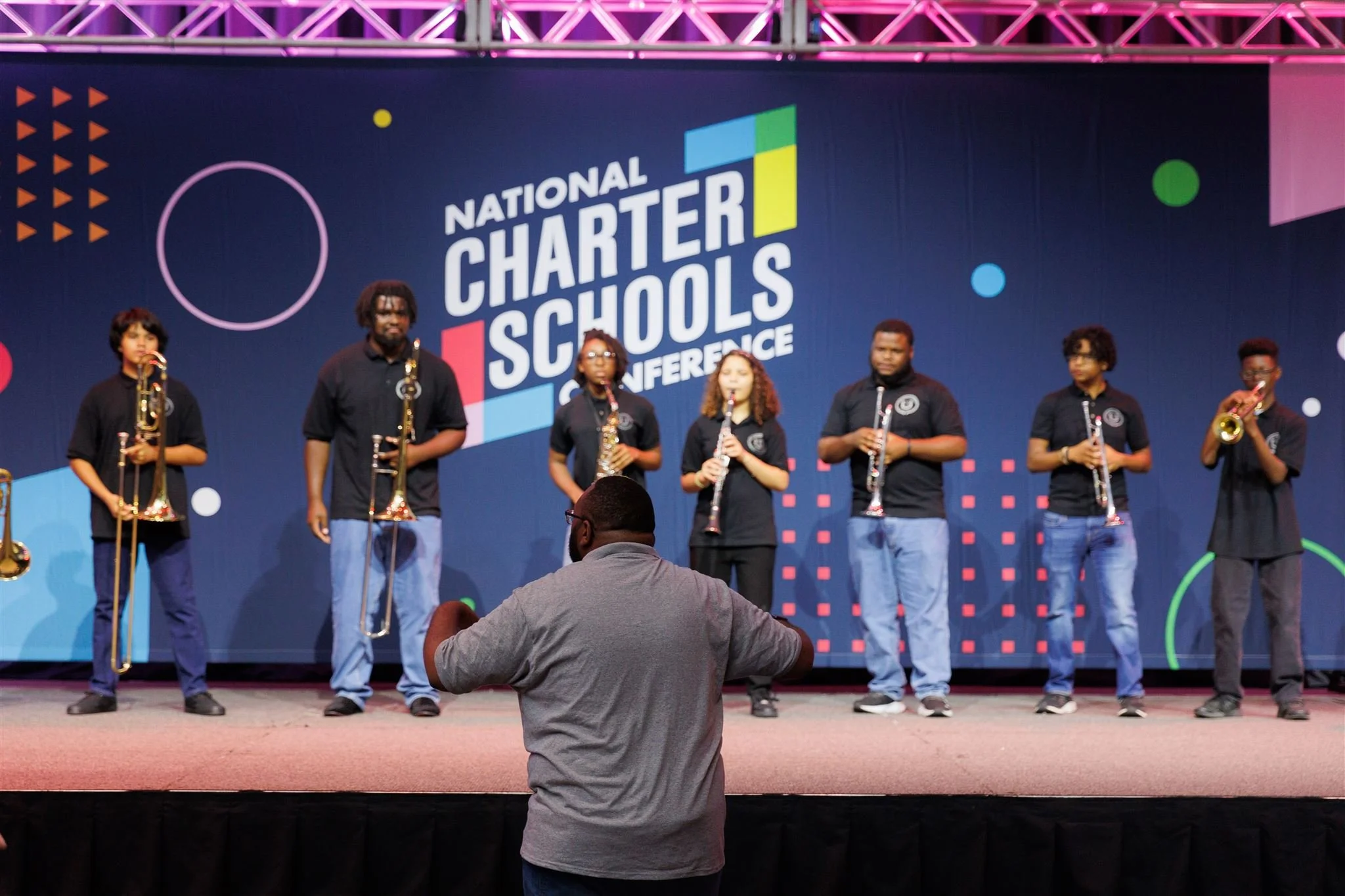 A group of seven students standing on a stage, holding musical instruments, during the National Charter Schools Conference.