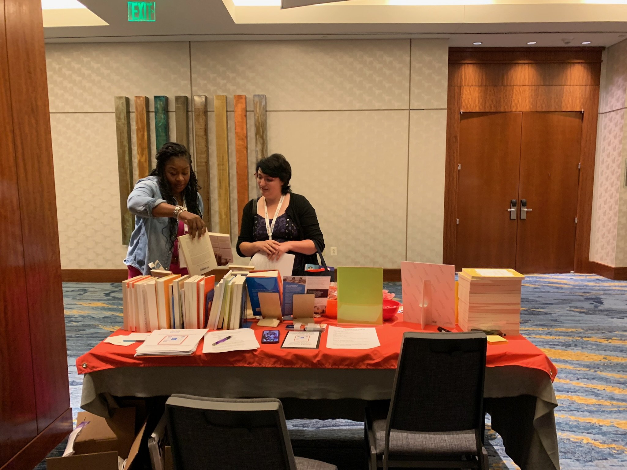Two women browsing books at a table display in a conference room. The table has multiple stacks of books and papers, with a red tablecloth. The room has patterned carpeting and beige walls with decorative wood panels and artwork.