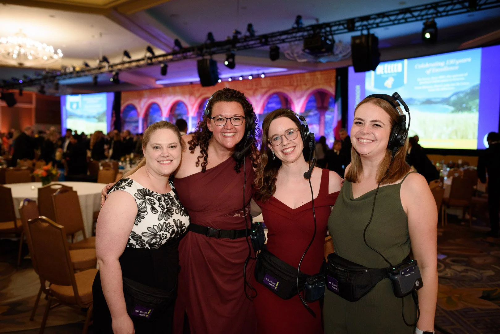 Four women at a conference, wearing headsets and name tags, standing together in a hotel ballroom with a stage and large screen in the background.