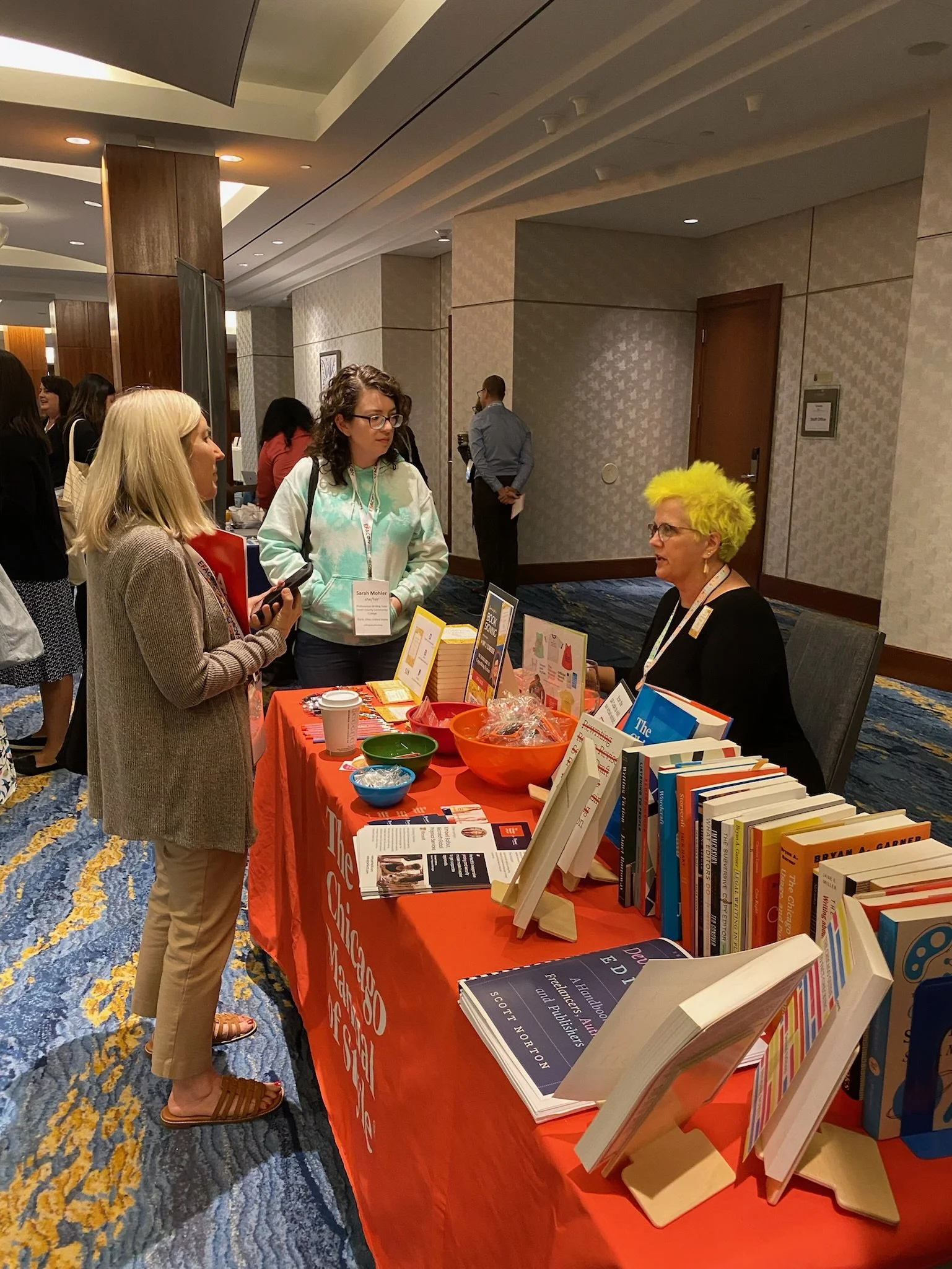 At a conference booth with a red tablecloth, a woman with bright yellow hair is conversing with two women. The table displays books, informational flyers, and bowls with giveaways or snacks.