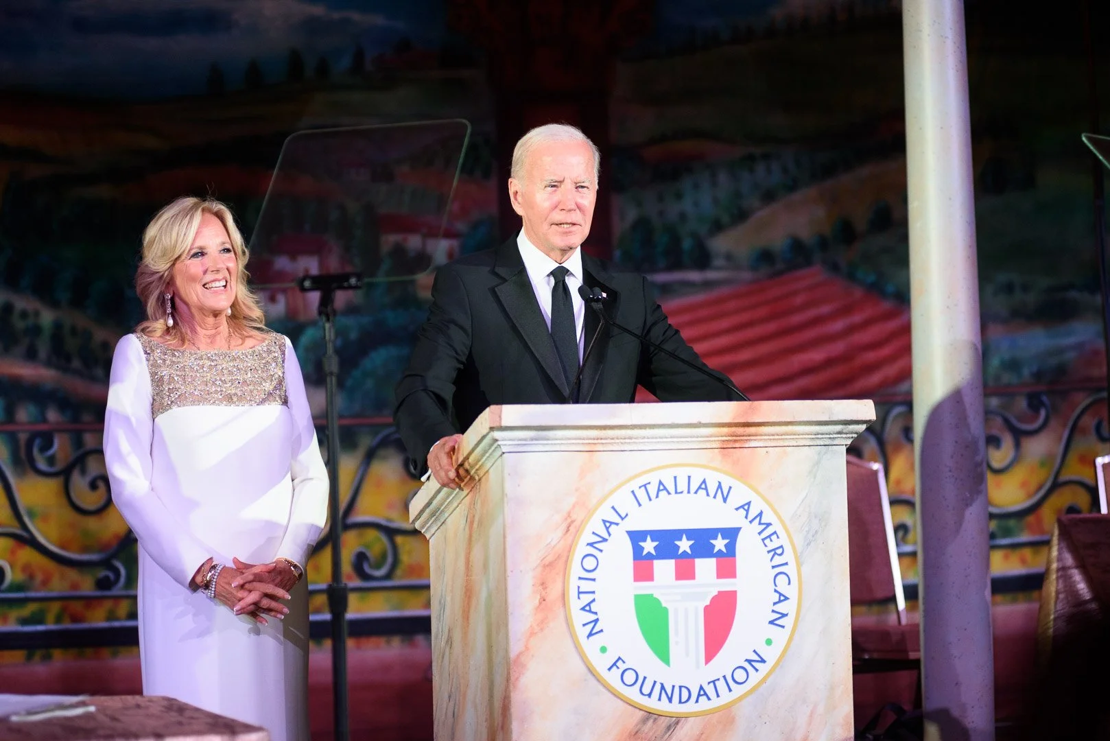 President Joe Biden and a woman standing at a podium with the National Italian American Foundation logo during an event, with a colorful backdrop.