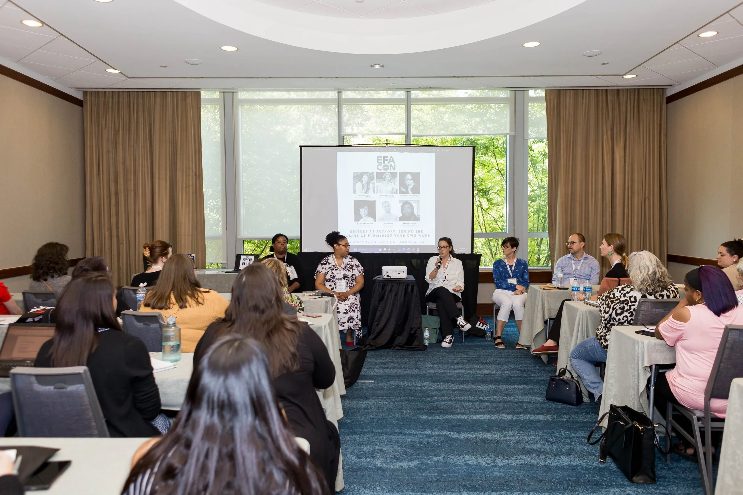 A panel discussion at a conference with eight women and two men seated on stage, in front of a large screen displaying a poster for the EFA Conference, in a room with large windows and beige curtains.