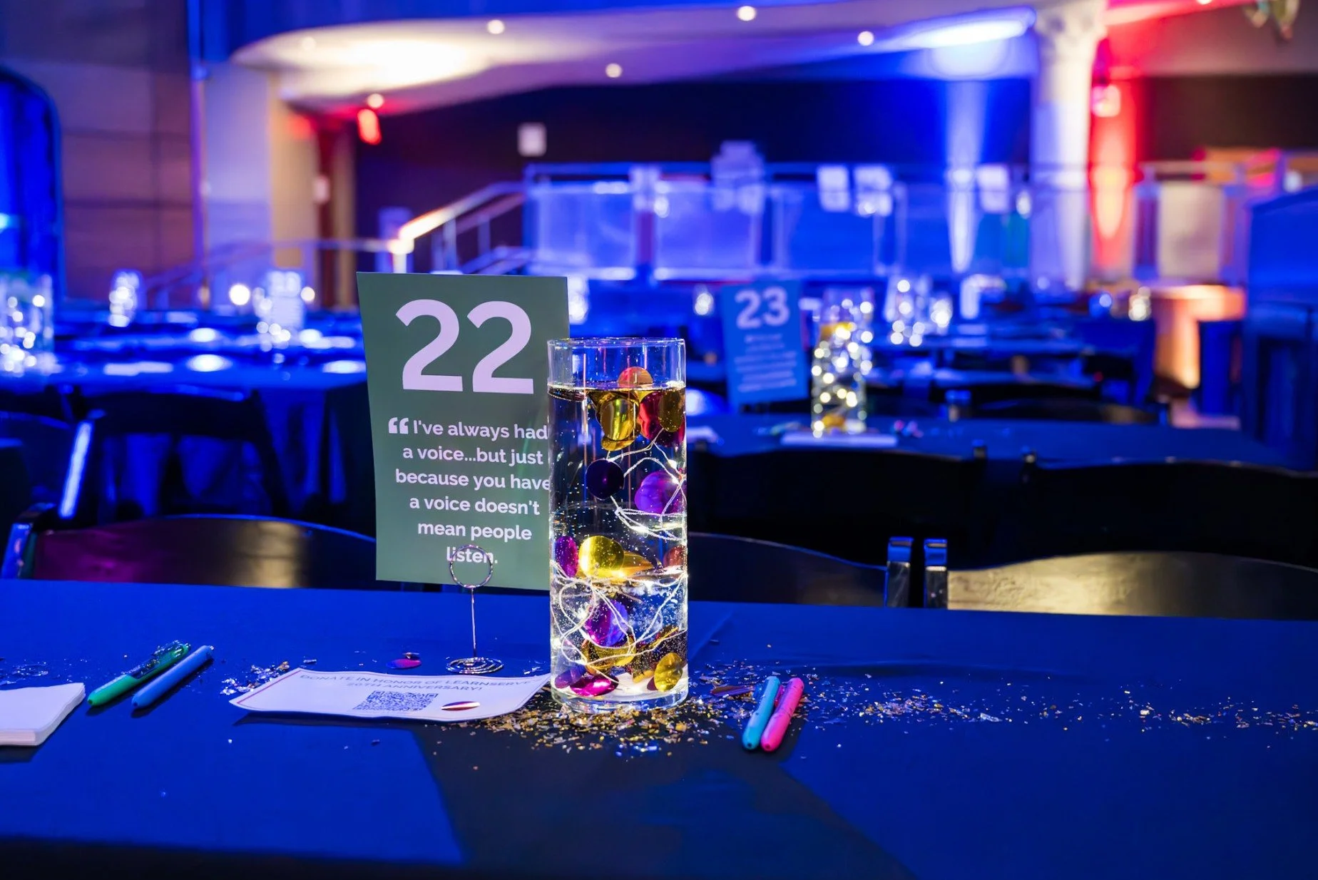 Table number 22 at a conference or event, with a transparent vase holding colorful decorative balls and string lights, surrounded by pens and a QR code on paper, in a dimly lit room with blue and red lighting.