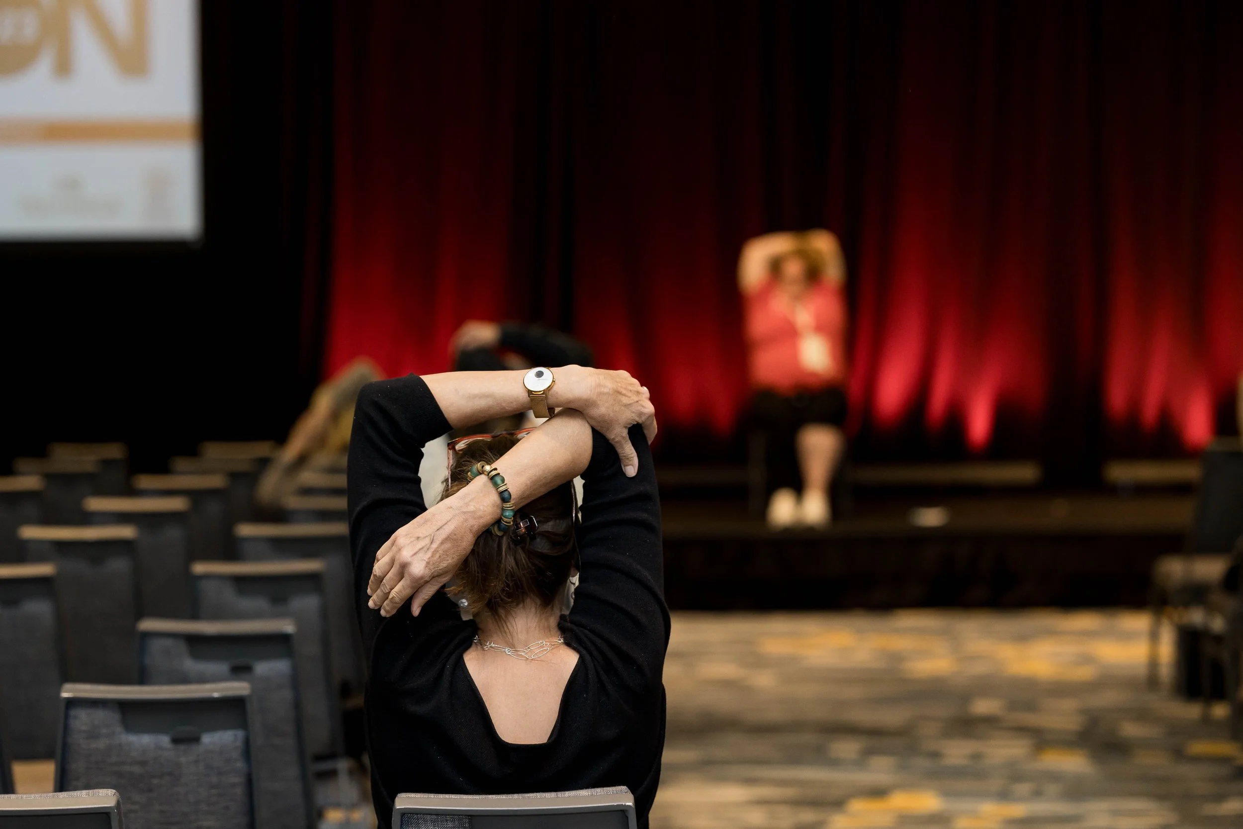 A woman seated in an empty auditorium stretching her arms behind her head, with a stage and red curtain in the background.