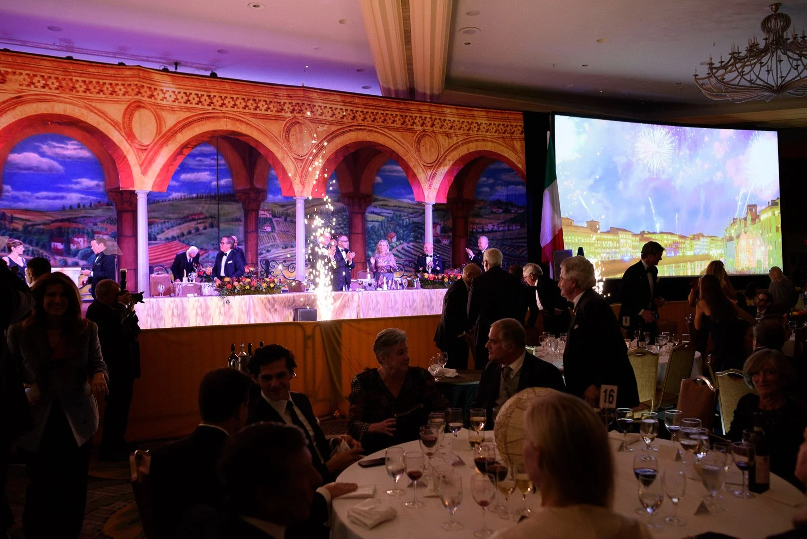 Elegant banquet hall with a stage decorated with a Venetian landscape mural, fireworks display on a large screen, and people in formal attire socializing and celebrating with fireworks and champagne.