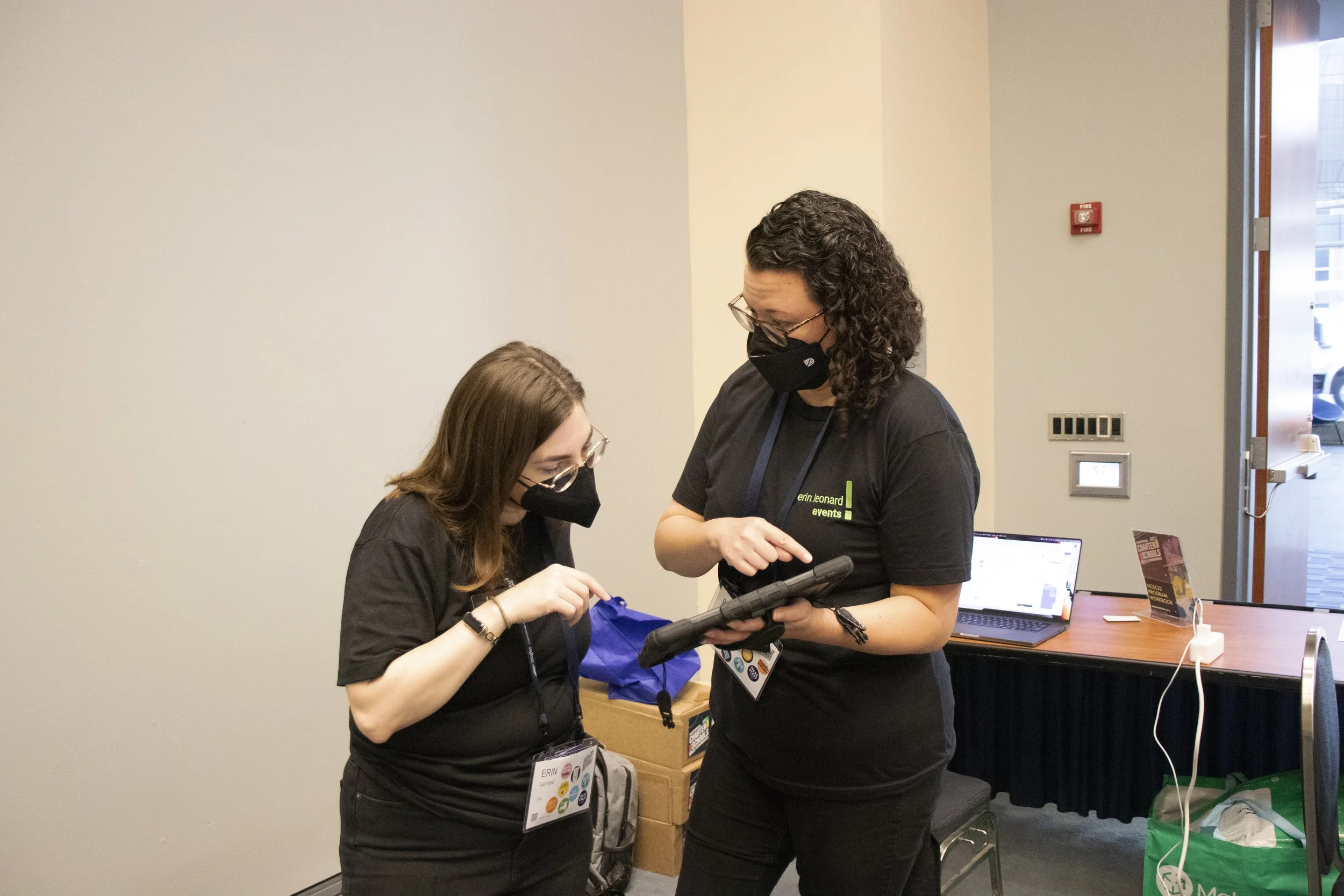 Two women wearing black shirts and face masks are looking at a tablet device together in a conference room. One woman has a name tag that reads 'Erin' and a lanyard with event badges.
