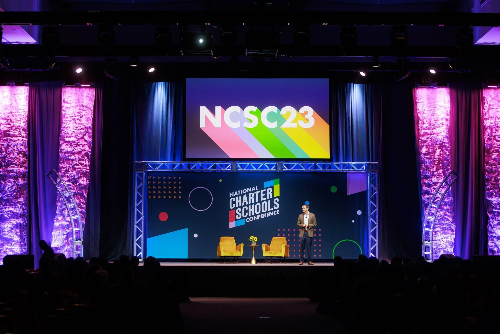 A man stands on stage at the National Charter Schools Conference, with colorful graphics and a large screen displaying NCSS23 and the conference title.