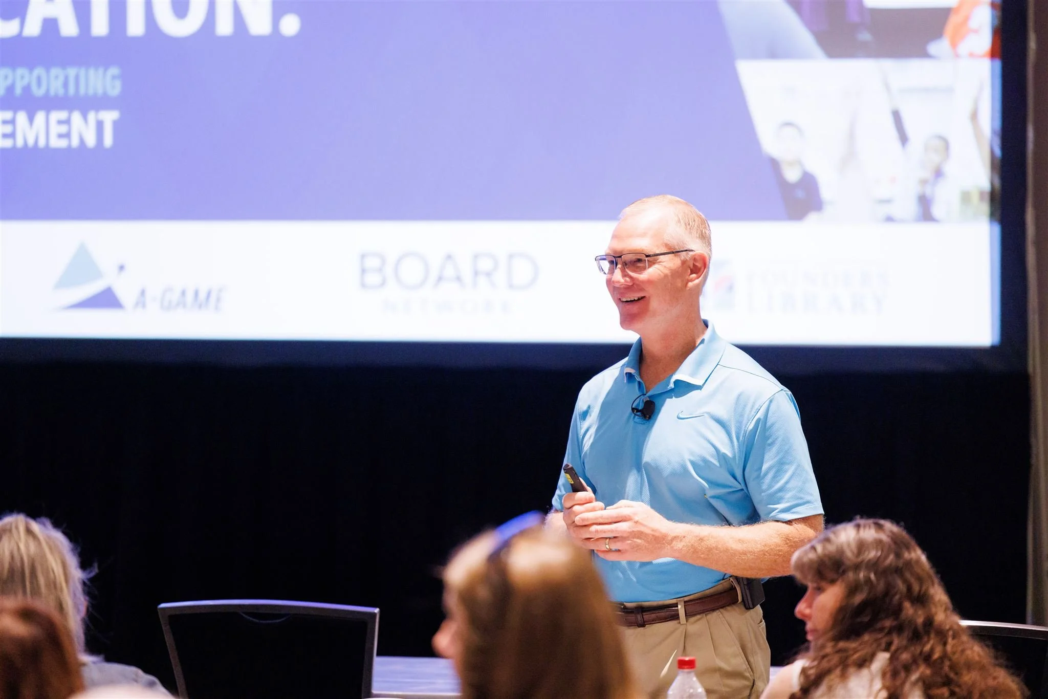 A man in glasses and a light blue shirt smiling and holding a remote control, standing in front of a large presentation screen during a conference.