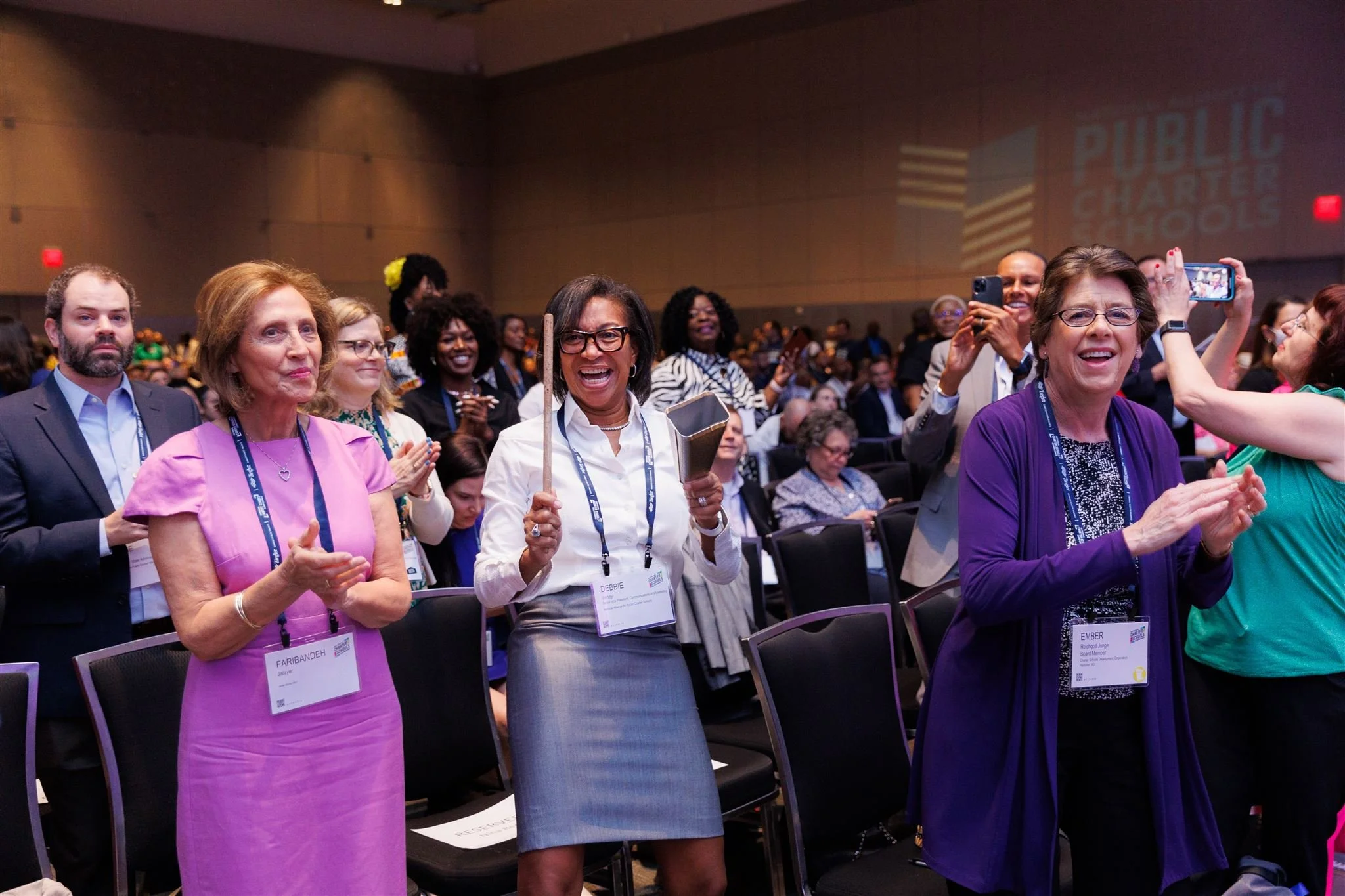 Group of professionals at a conference, some clapping and cheering, with a projected logo for 'Public Charter Schools' on the wall in the background.