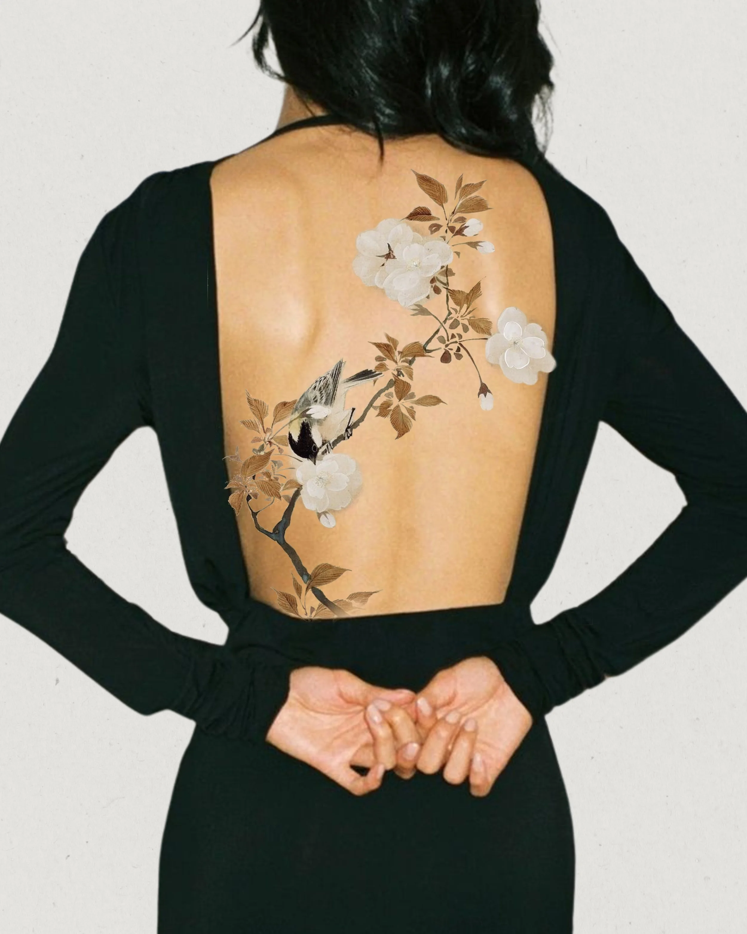 Back tattoo of a bird on a flowering branch on a woman's back, wearing a black dress with an open back.