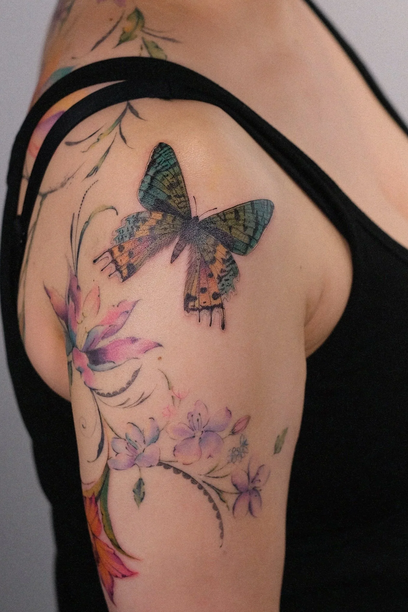 Close-up of a person's shoulder and upper arm with a colorful butterfly tattoo surrounded by floral designs.