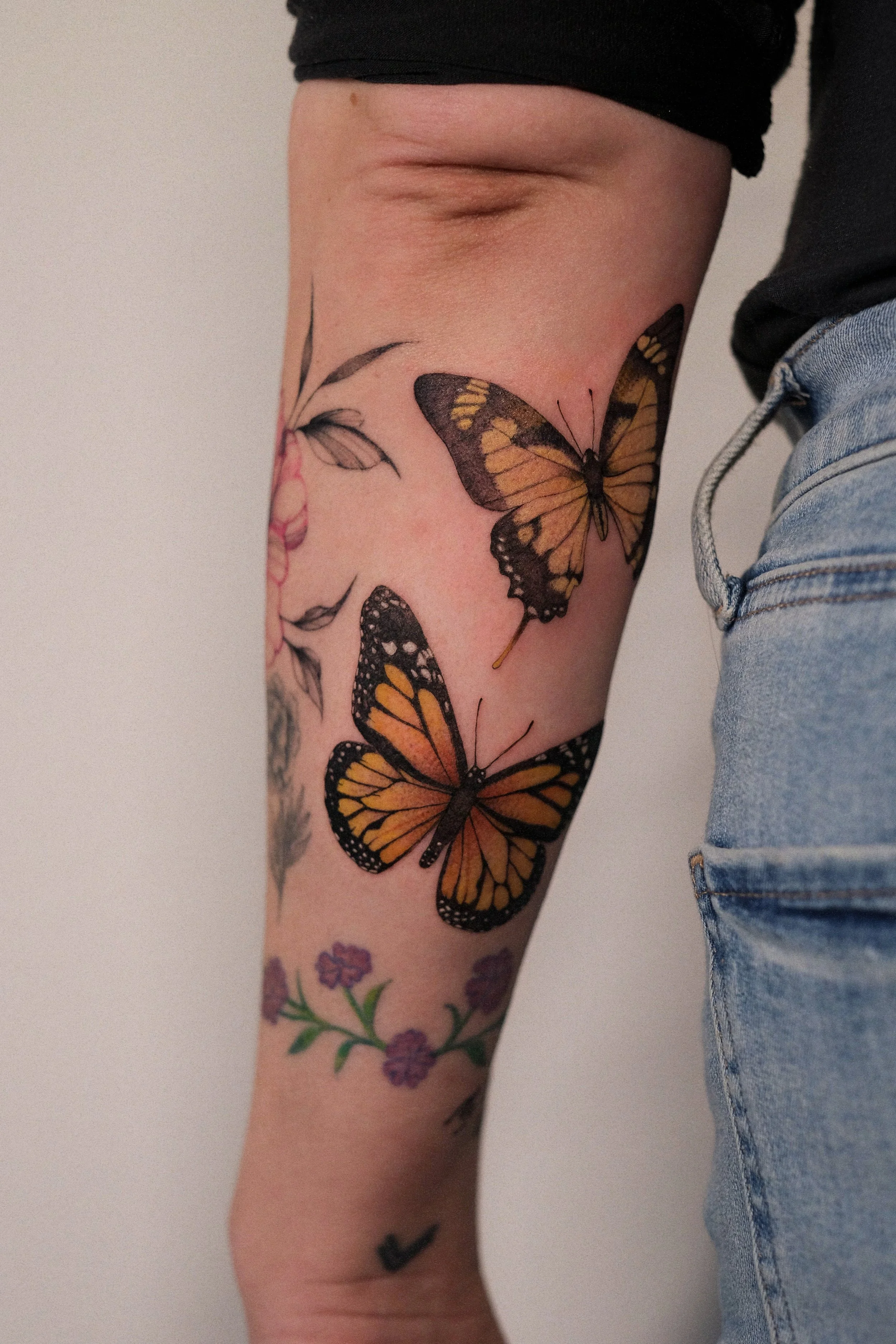 Close-up of a person's forearm with multiple colorful tattoos, including butterflies and flowers, wearing a black shirt and light blue jeans.