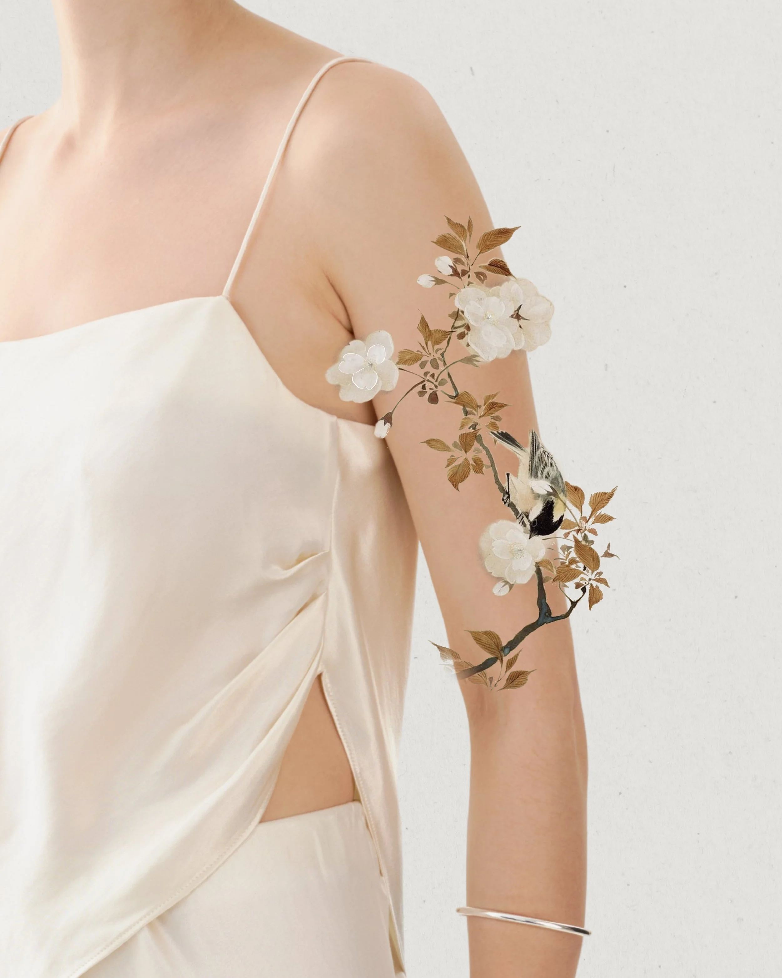 A woman in a cream-colored satin top with spaghetti straps, showcasing a floral tattoo on her upper arm.