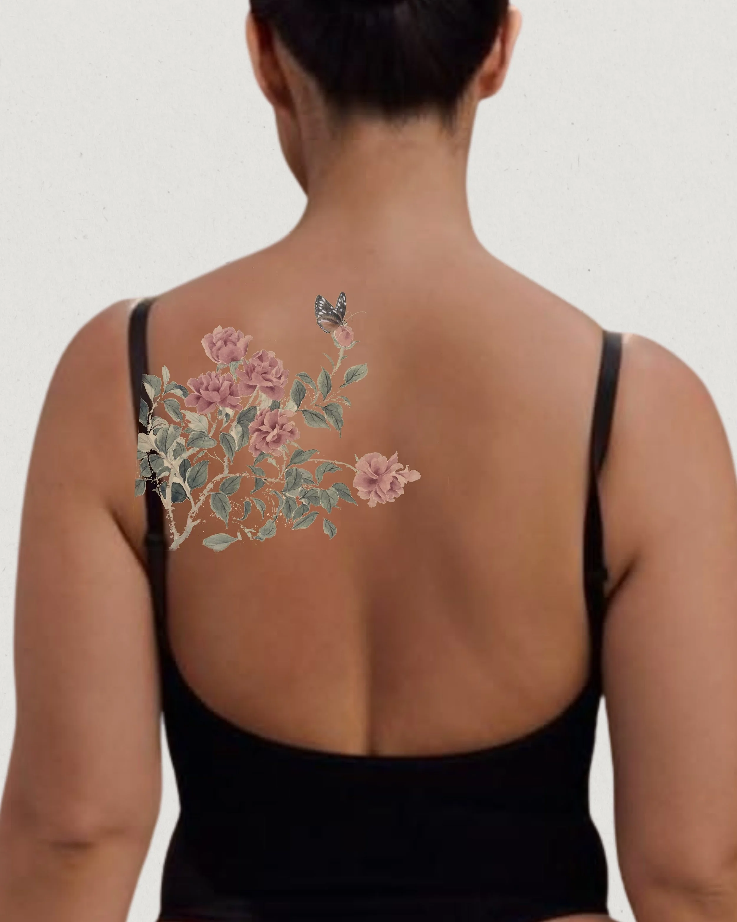 A woman with short dark hair, wearing a black spaghetti strap top, has a floral tattoo with pink flowers and green leaves on her upper back, with a butterfly perched on one of the flowers.