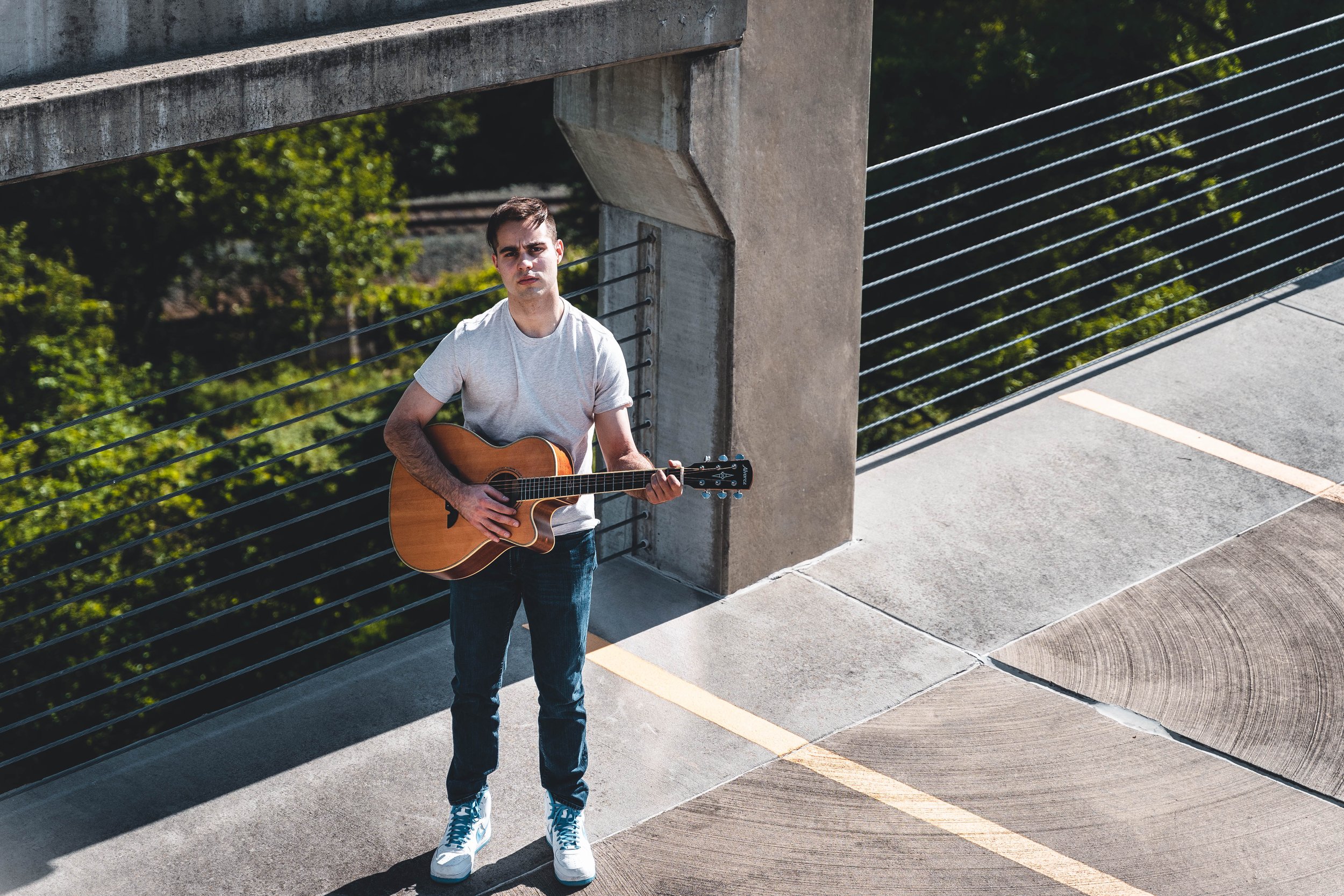 Young man in a gray t-shirt and jeans playing guitar outdoors on a sidewalk next to a concrete railing with greenery in the background.